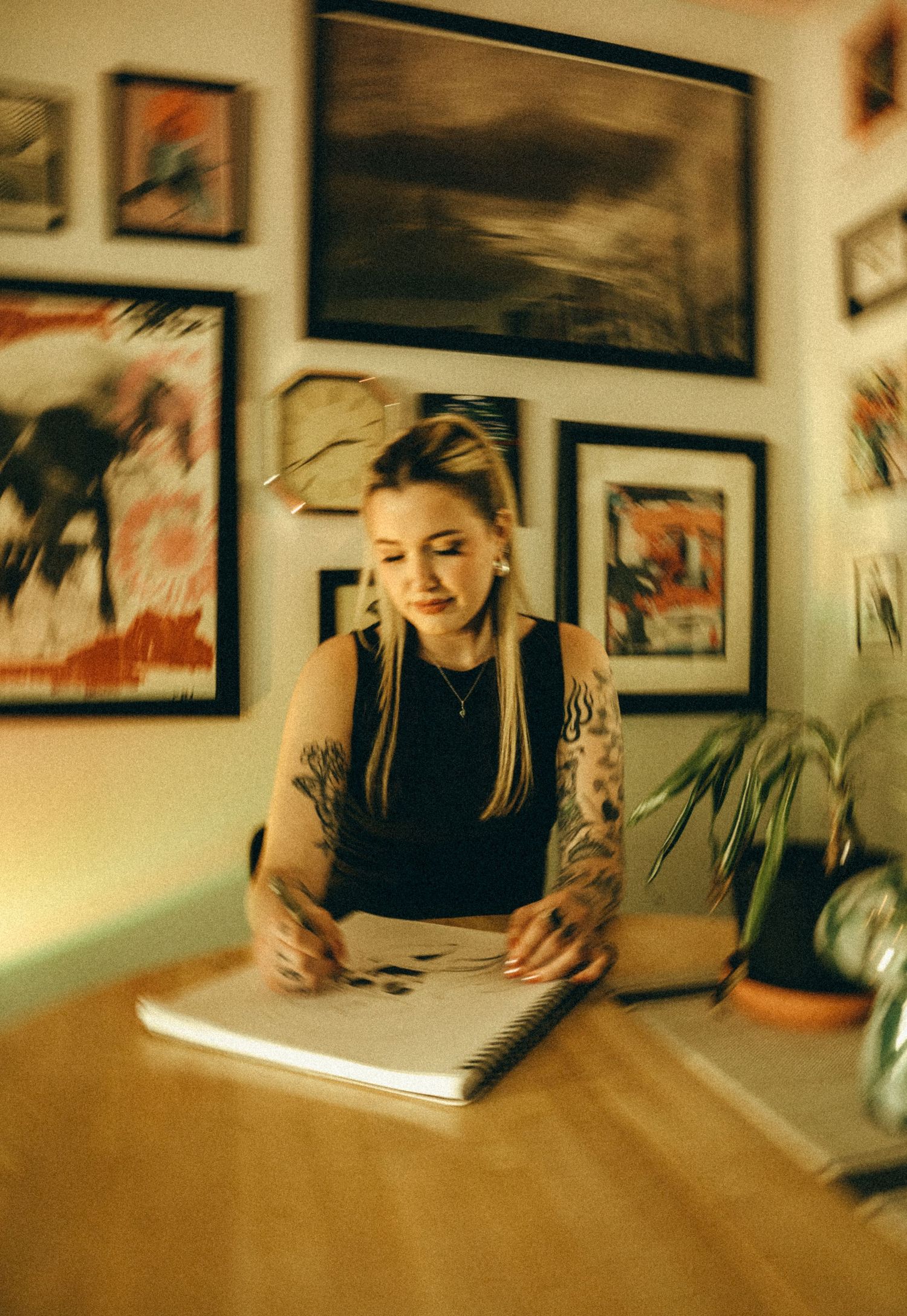 Tattooed blonde woman sketching in notebook at desk surrounded by framed artwork on warm walls.