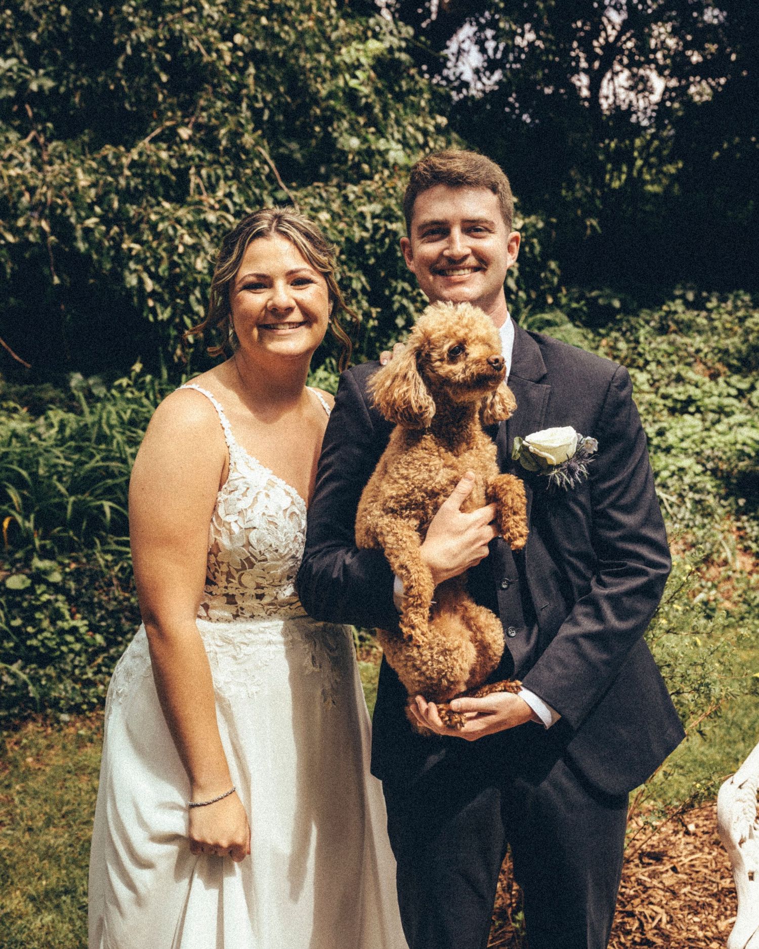 Smiling bride and groom holding a fluffy brown toy poodle in an outdoor garden setting.