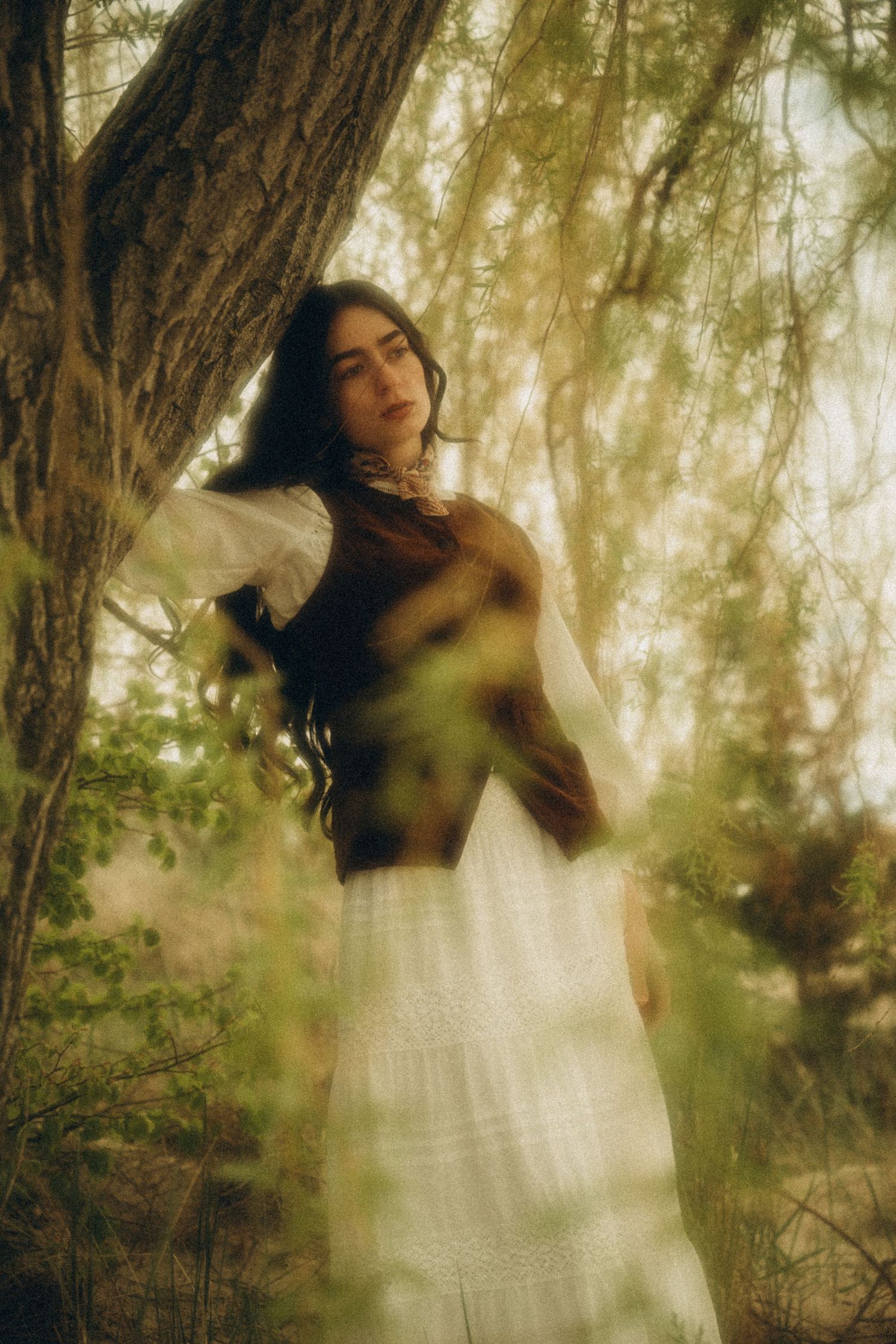 Dark-haired woman in white lace skirt leaning against tree in soft focus woodland portrait.
