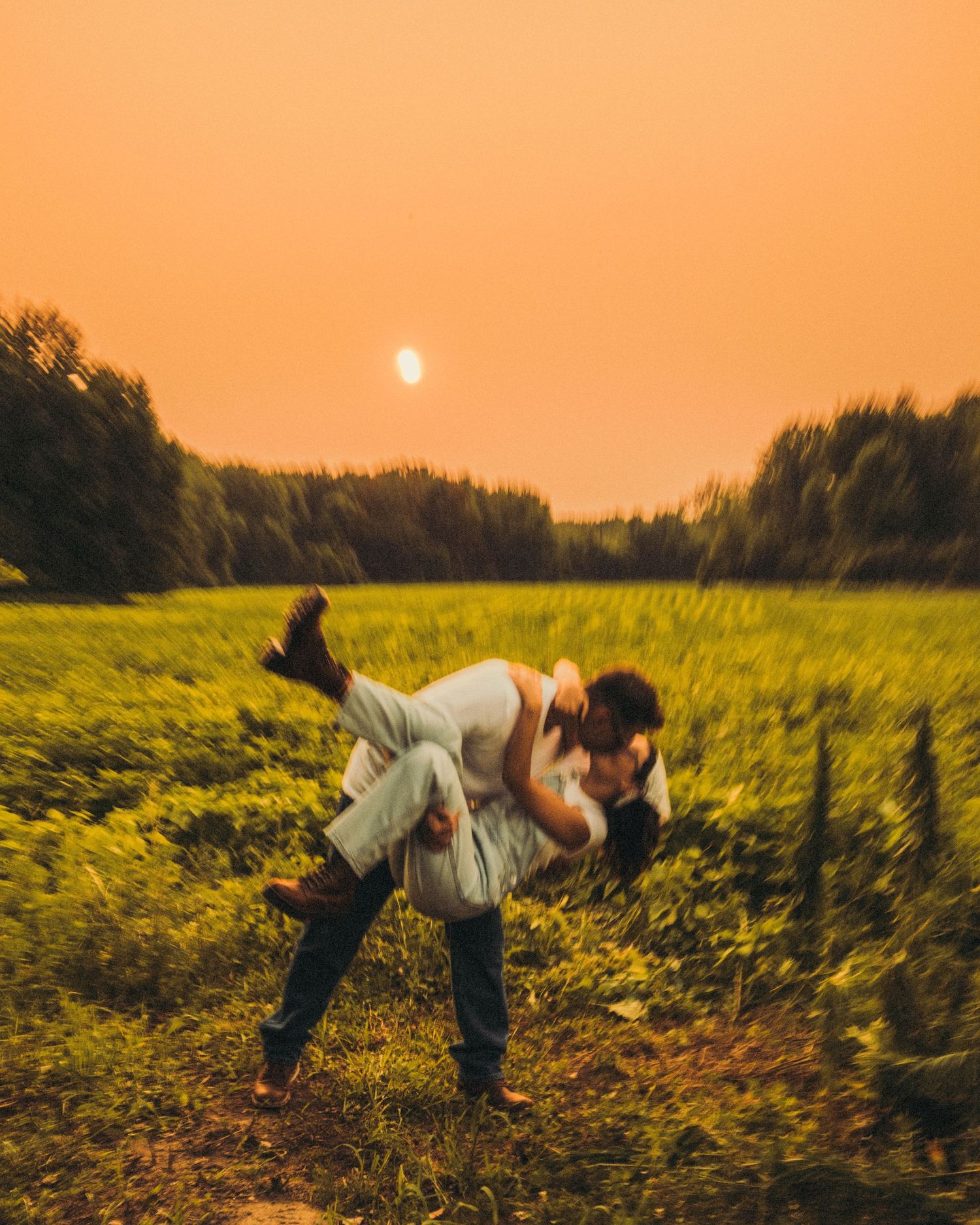 A man dips a woman in a green field under a dramatic orange sunset sky.