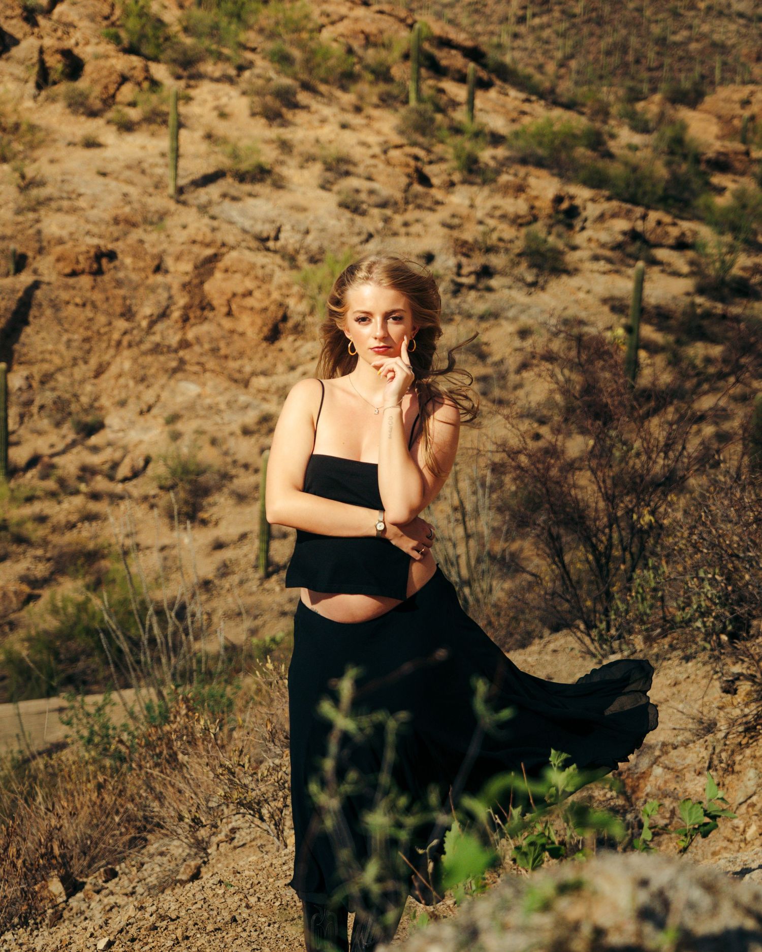 A blonde woman in a flowing black two-piece outfit poses on a desert hillside with cacti.