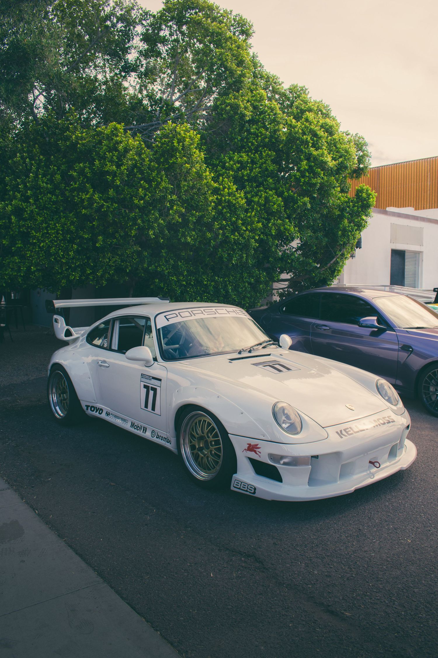 White Porsche 993 race car with number 77 and sponsor decals parked in front of green trees.