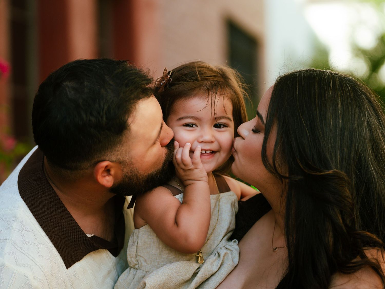 Two parents kissing their smiling toddler daughter on the cheeks in a sweet outdoor family portrait.