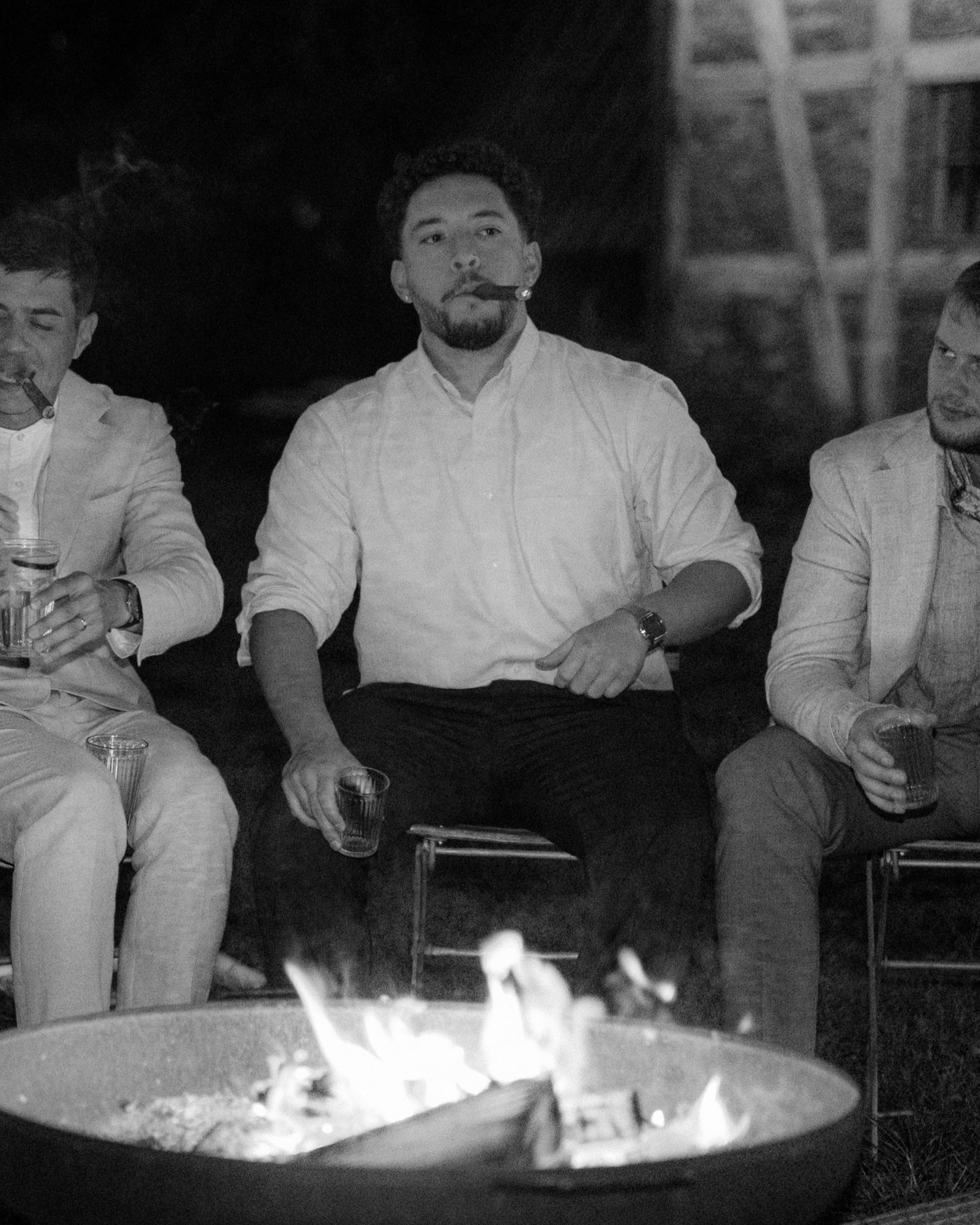Three men sitting around an outdoor fire pit at night, one smoking a cigar, in black and white photo.