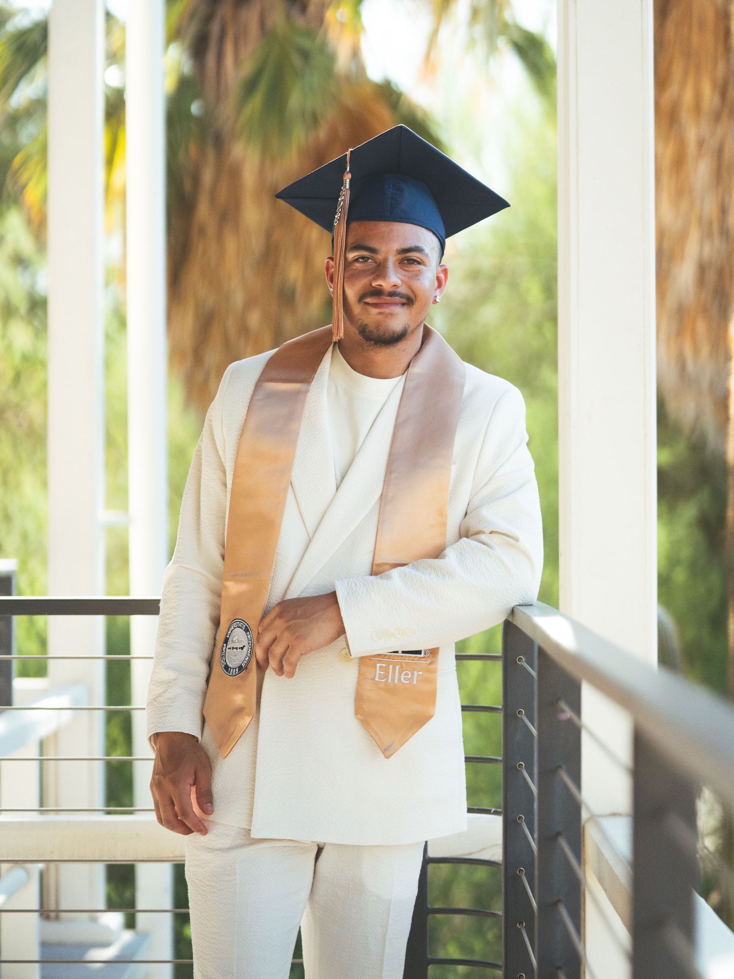 Young man in white suit and graduation cap smiling on a balcony surrounded by palm trees at the University of Arizona