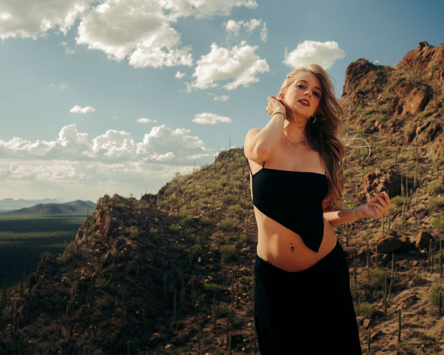 Woman in black off-shoulder dress posing on a rocky desert hillside under cloudy sky.