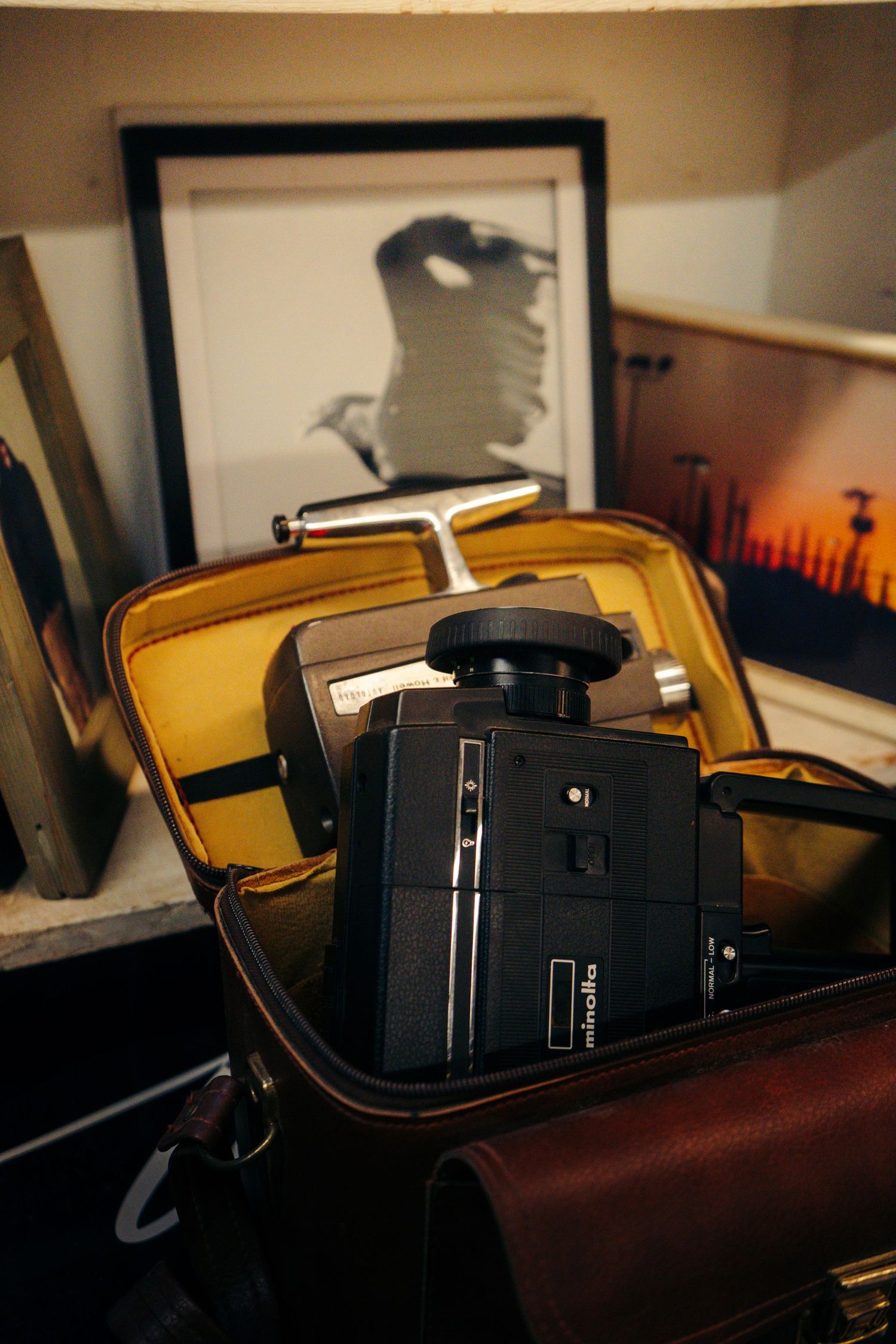 Vintage Olympus camera resting in a brown leather case beside framed photographs on a shelf.