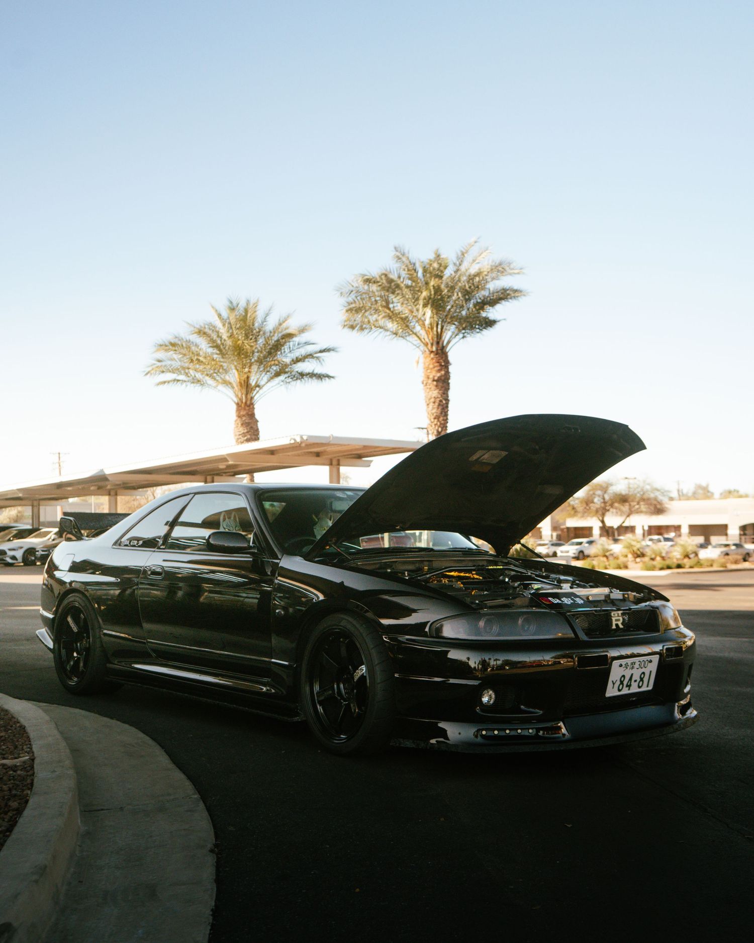 Black Nissan Skyline R33 with hood open parked near palm trees in a clear blue sky.