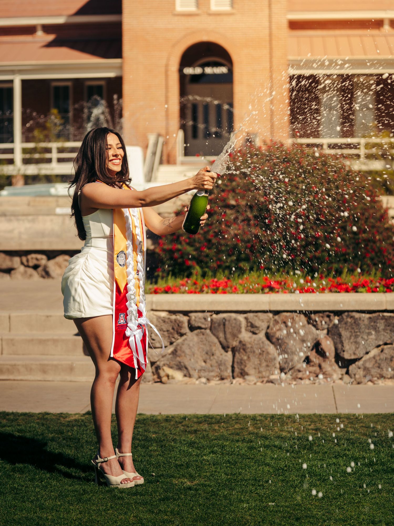 Smiling University of Arizona graduate in white dress popping champagne bottle on a sunny campus lawn.