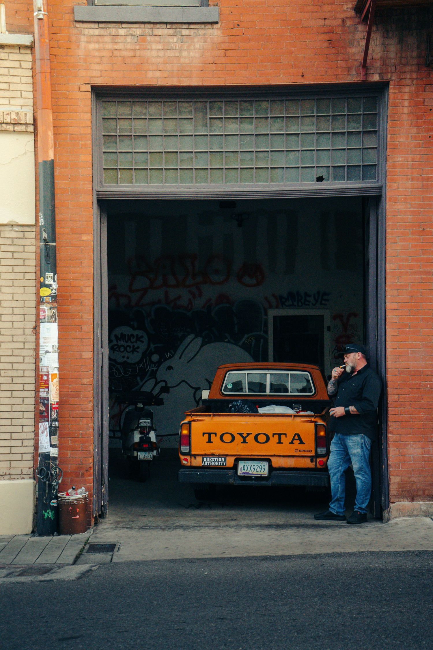 Man standing beside an orange Toyota pickup truck in a graffiti-covered urban garage doorway.