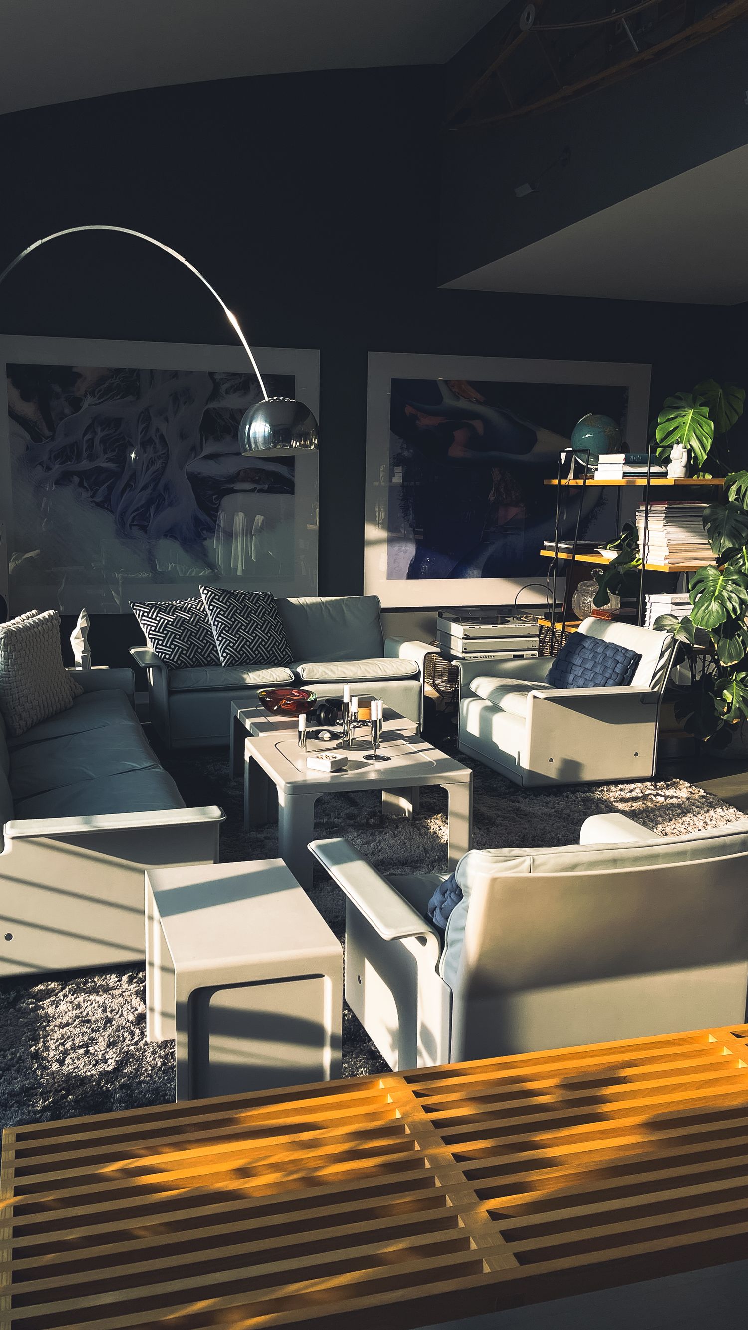 Sunlit modern living room with white furniture, arc lamp, and dark walls featuring large artwork.