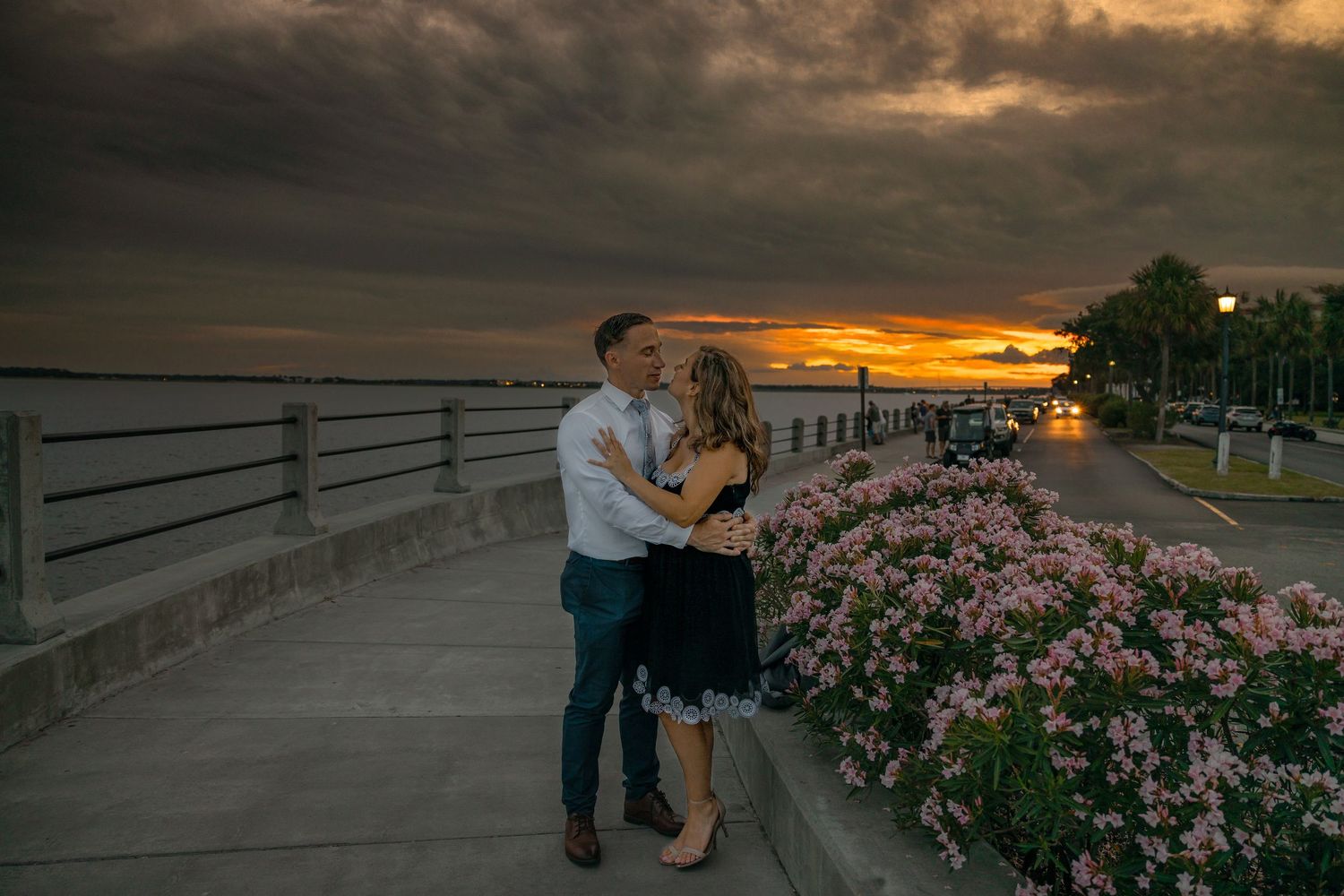 Dramatic orange sunset sky over waterfront boardwalk with pink flowering plants.