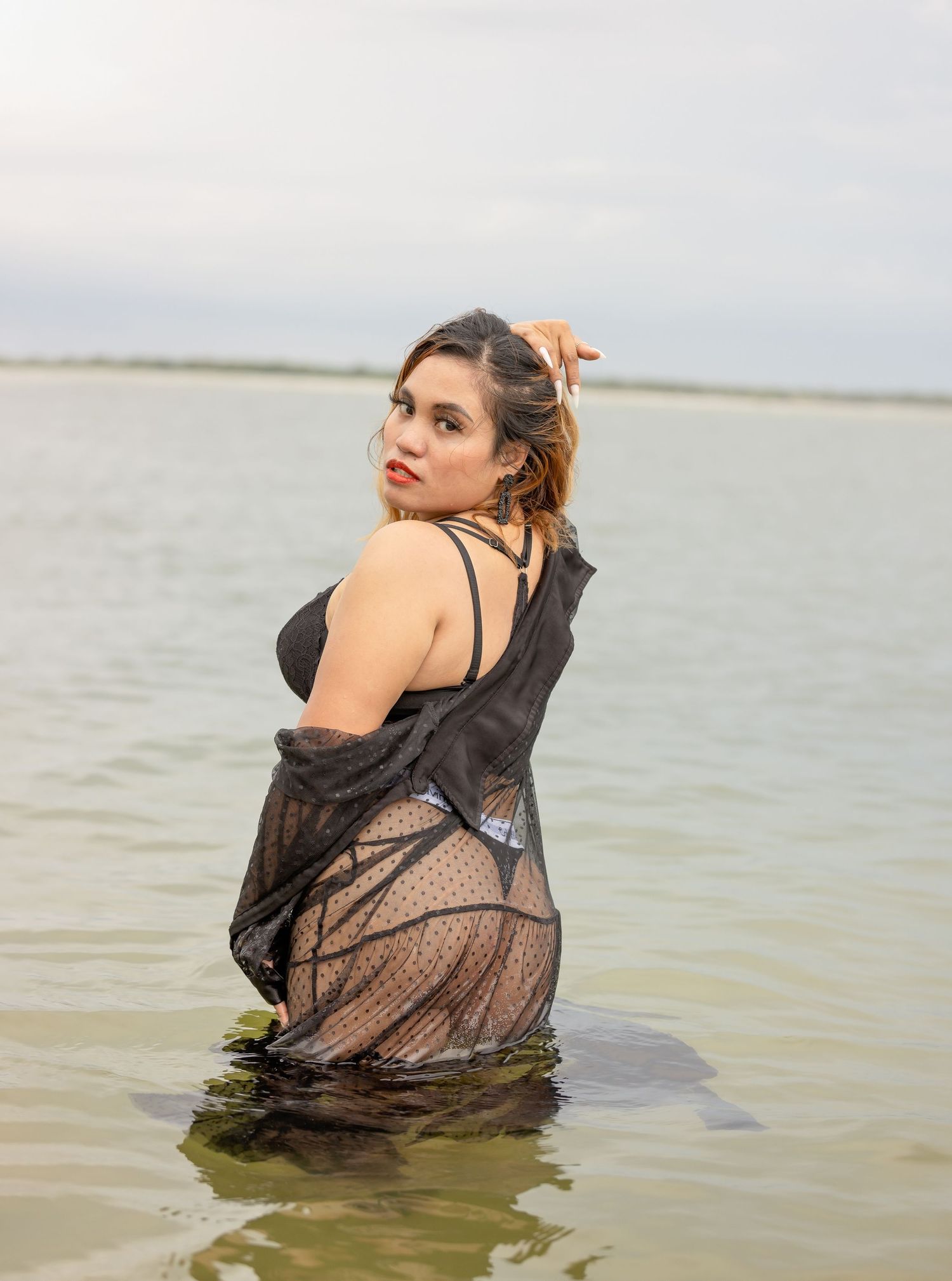 Woman in black mesh swimsuit cover poses in shallow water during beach photoshoot.