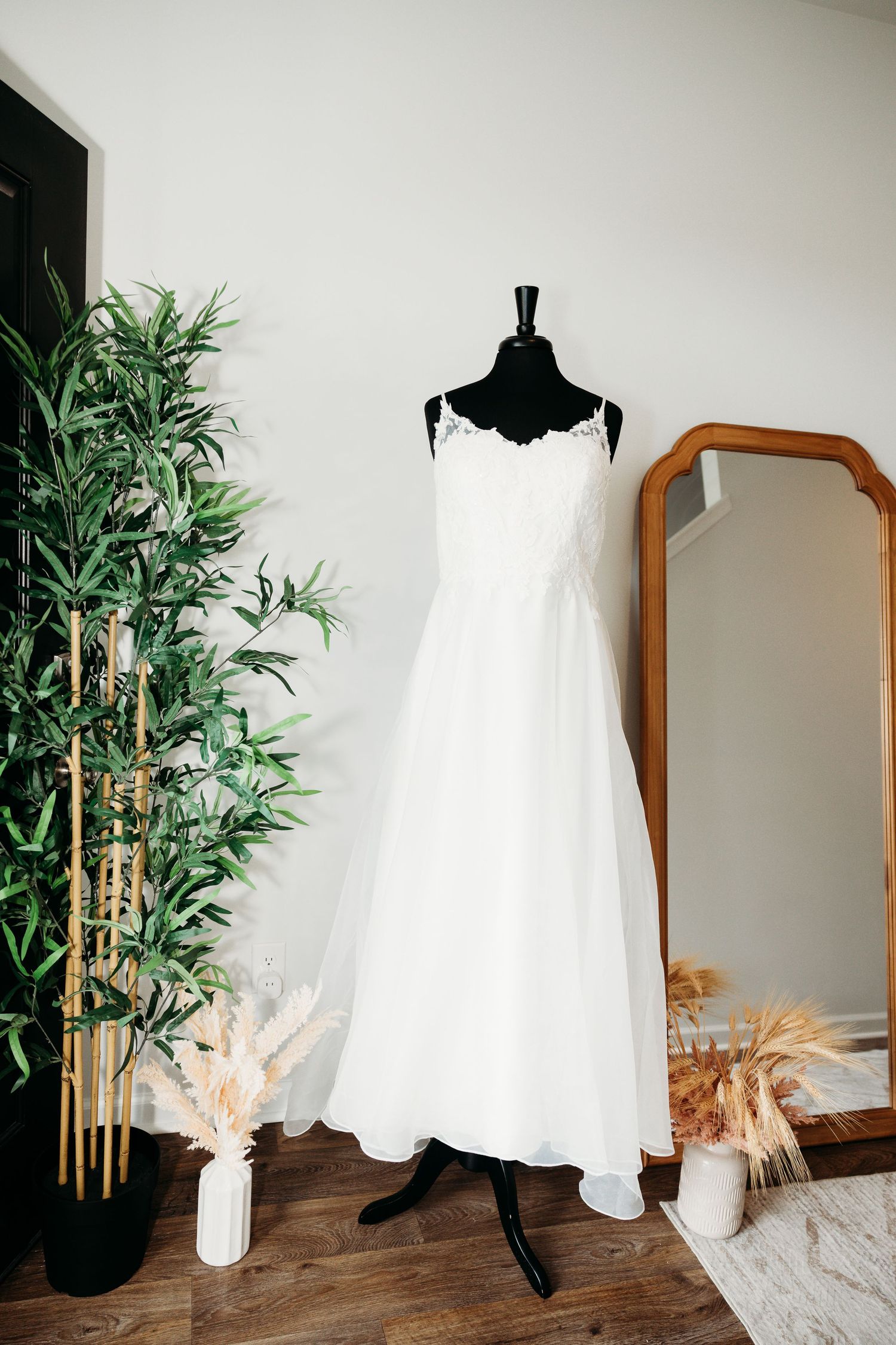 White wedding dress displayed on black mannequin with bamboo plants and wooden mirror in background.