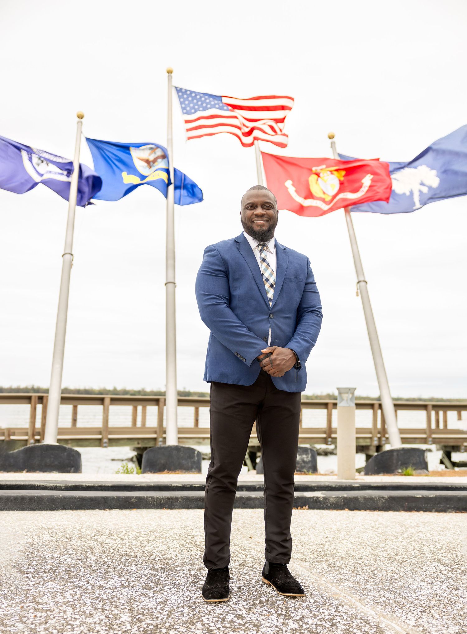 Person in navy suit stands near flagpoles with American and military flags waving in background.