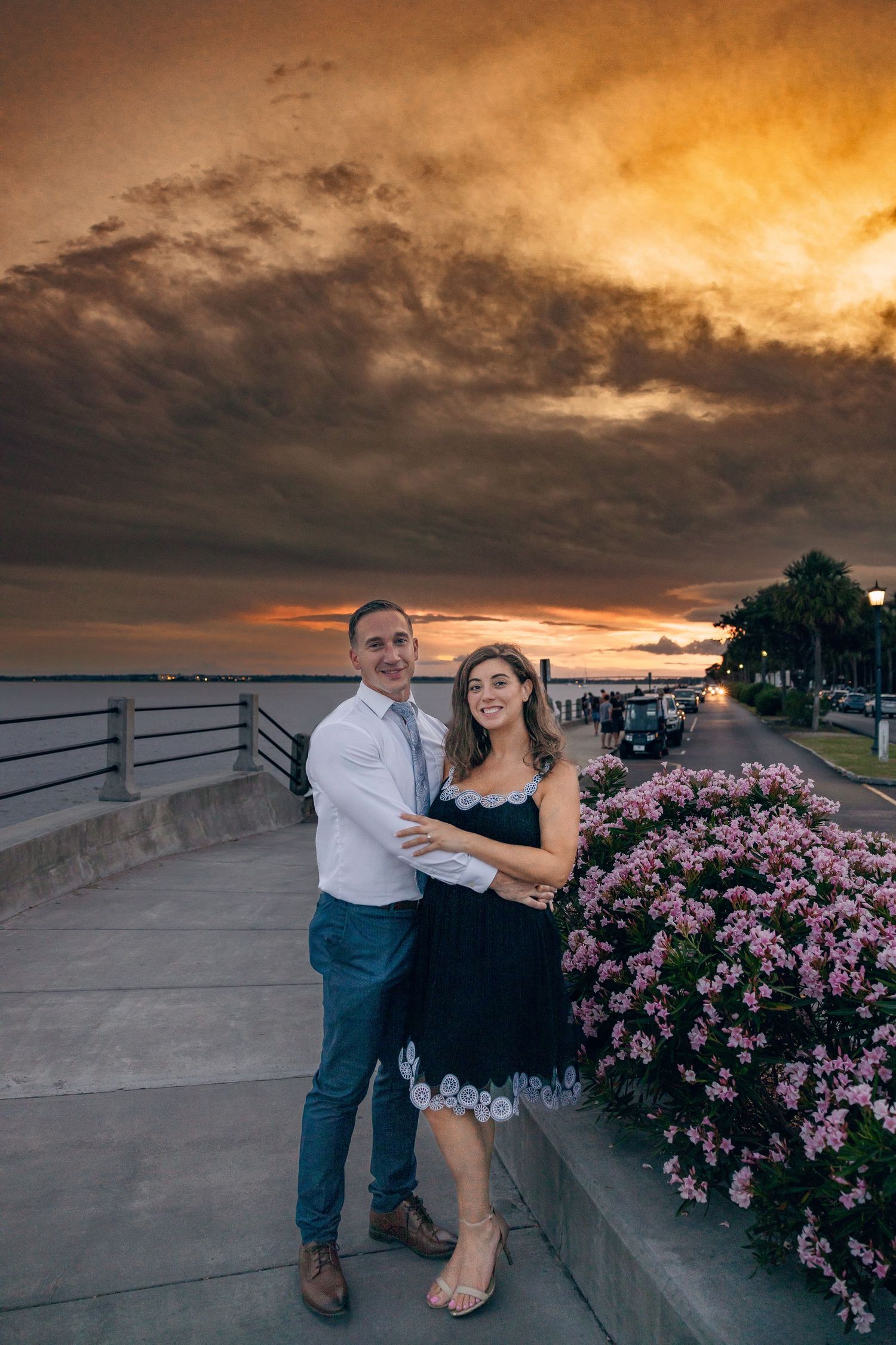 Couple sits together on concrete wall during dramatic sunset with pink flowers nearby.