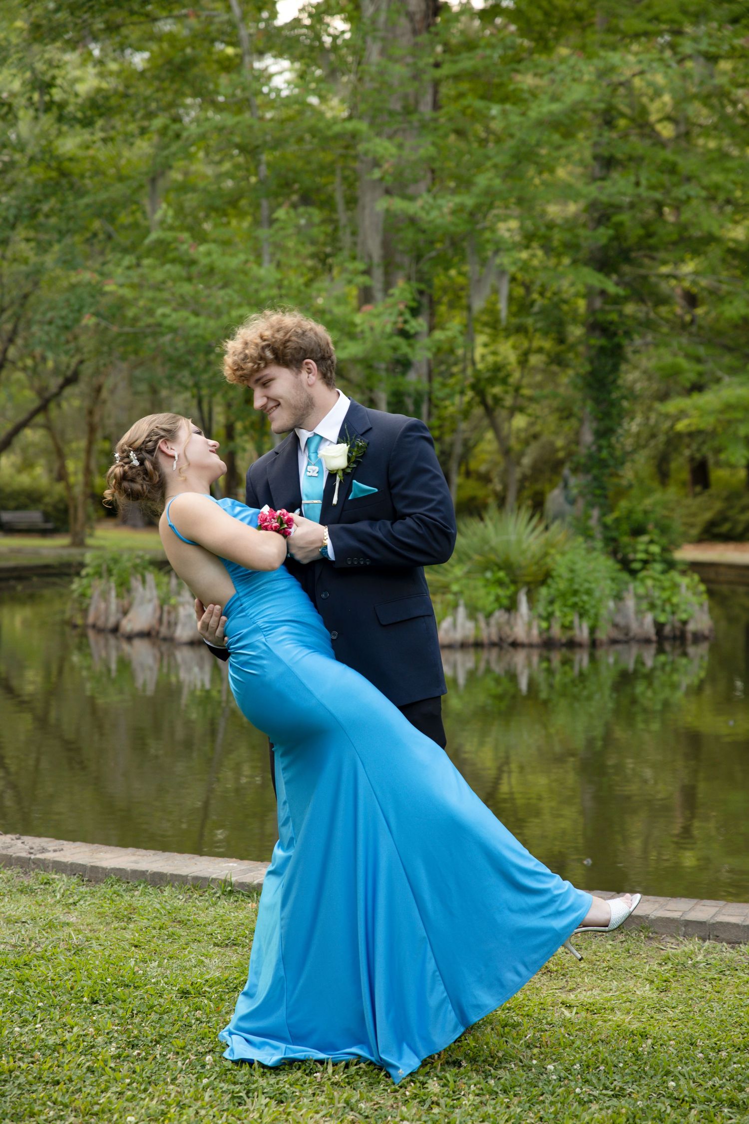 Dancing couple in blue dress and black suit pose dramatically on grass near water.