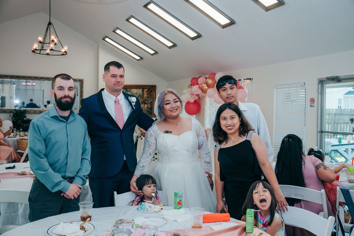Wedding reception celebration with guests gathered indoors around decorated tables and balloons.