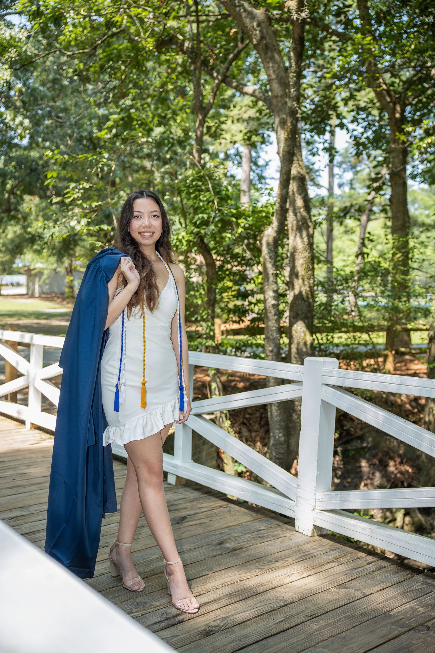 A graduate in white dress poses on a wooden bridge in a park setting.