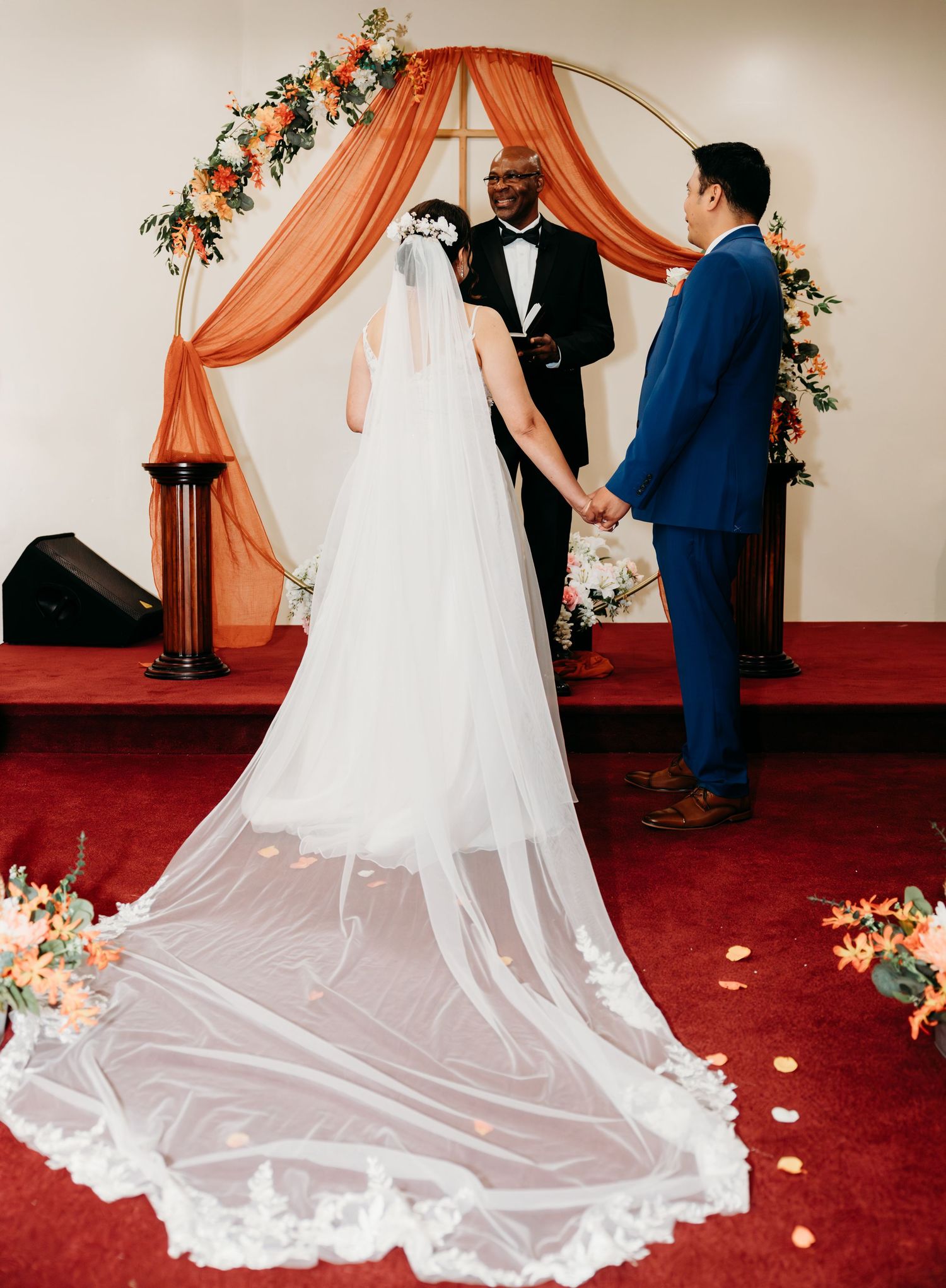 Wedding couple stands under a draped coral and floral arch during their indoor ceremony.