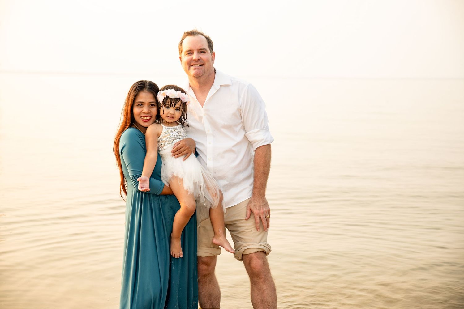 Family wearing coordinated white and teal outfits smiles together on beach at sunset.