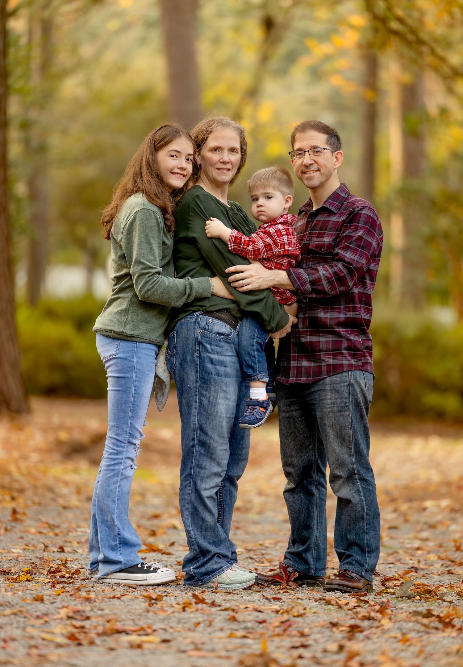 Family members wearing plaid and denim embrace while standing on fallen autumn leaves in a forest setting.