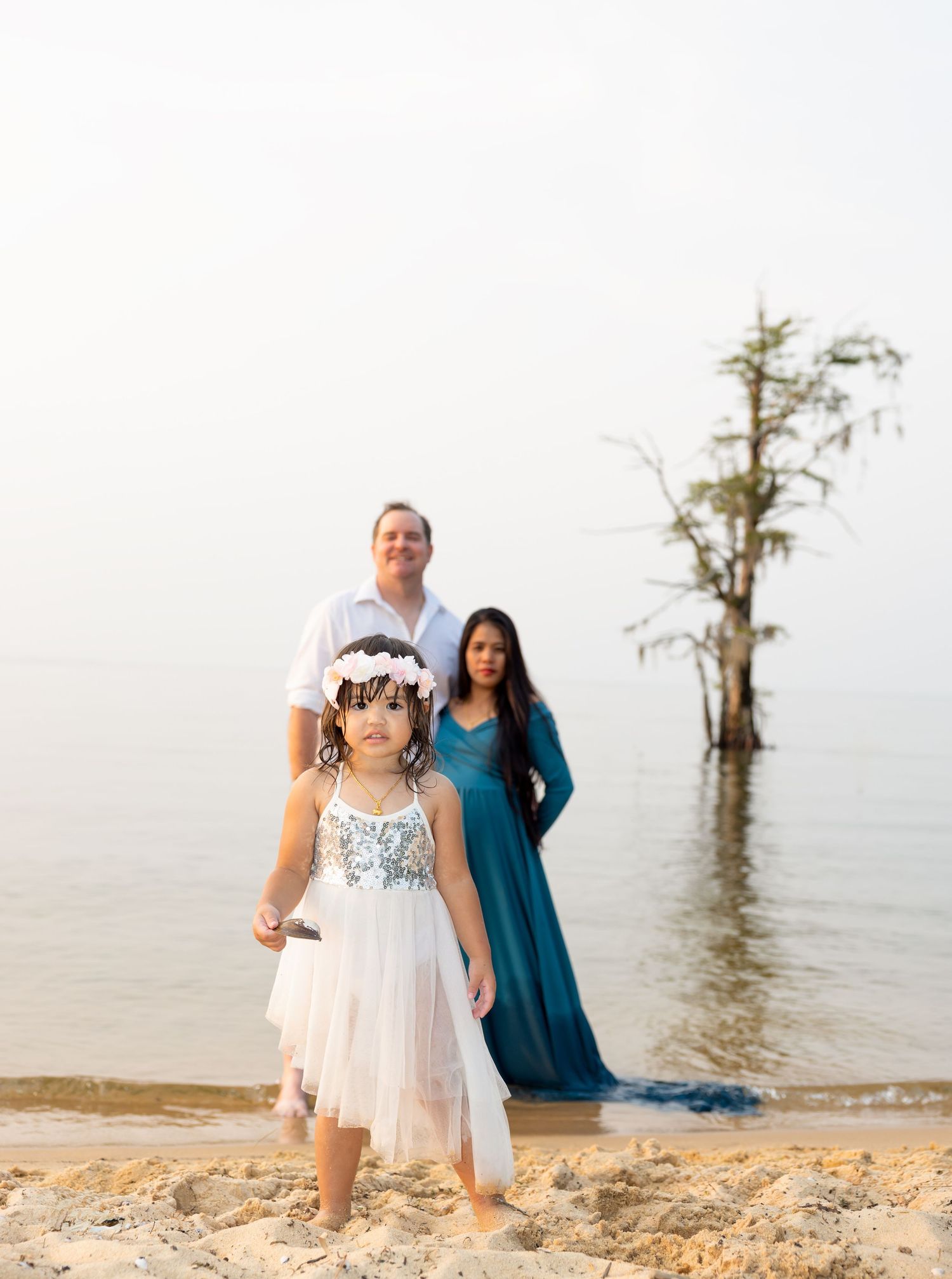 Family in white and blue outfits poses together on beach with cypress trees in background.