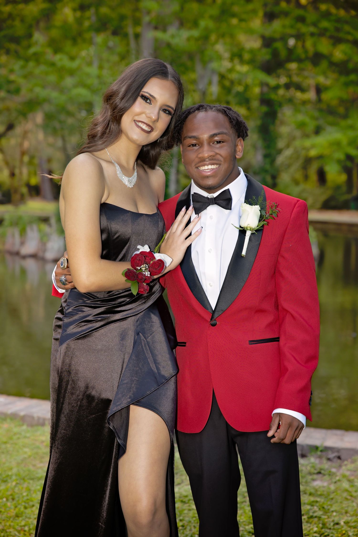 Formal couple poses together outdoors, wearing black dress and red tuxedo jacket.