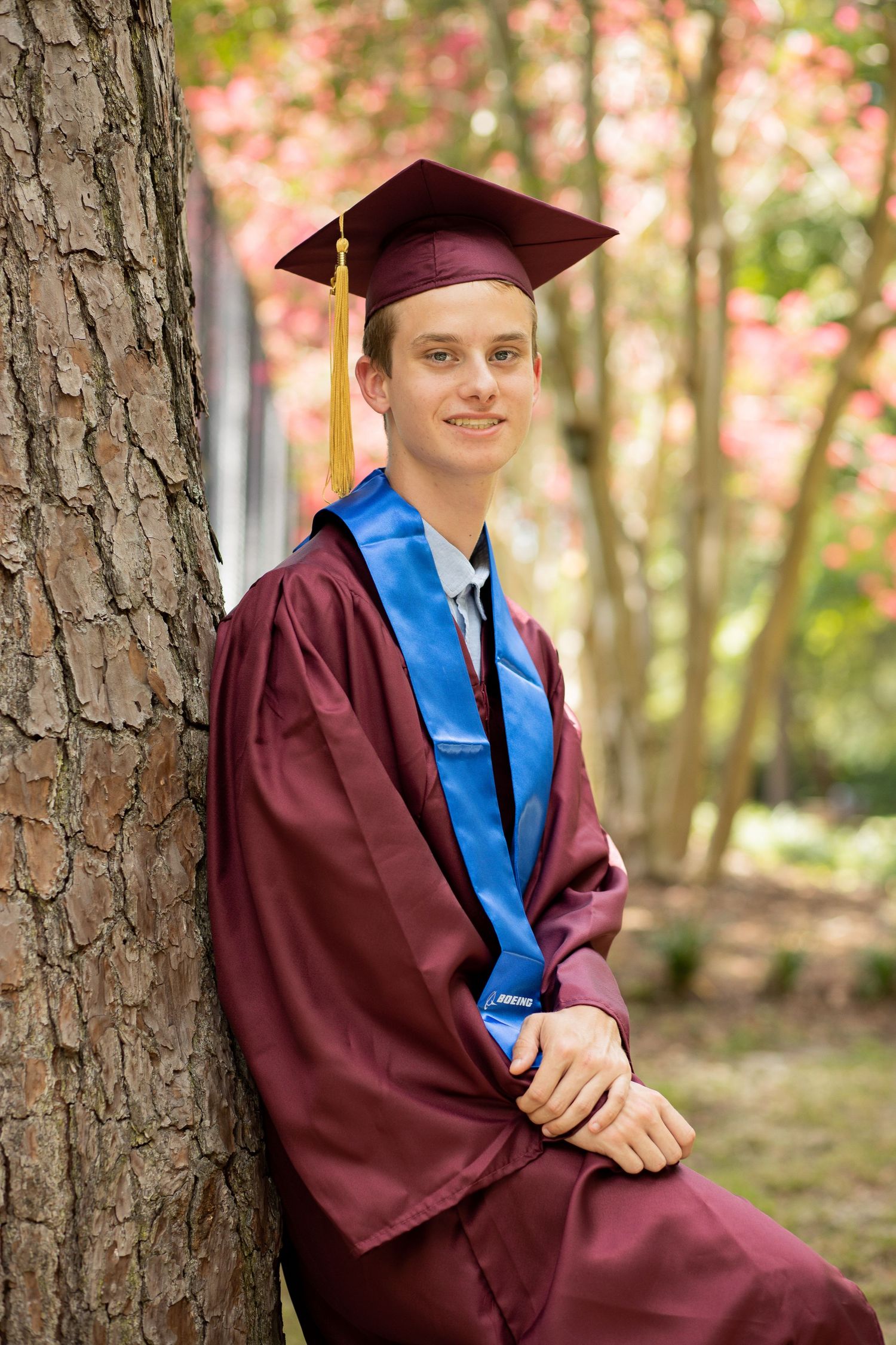 Graduate in maroon cap and gown with blue stole poses against trees in outdoor portrait session.