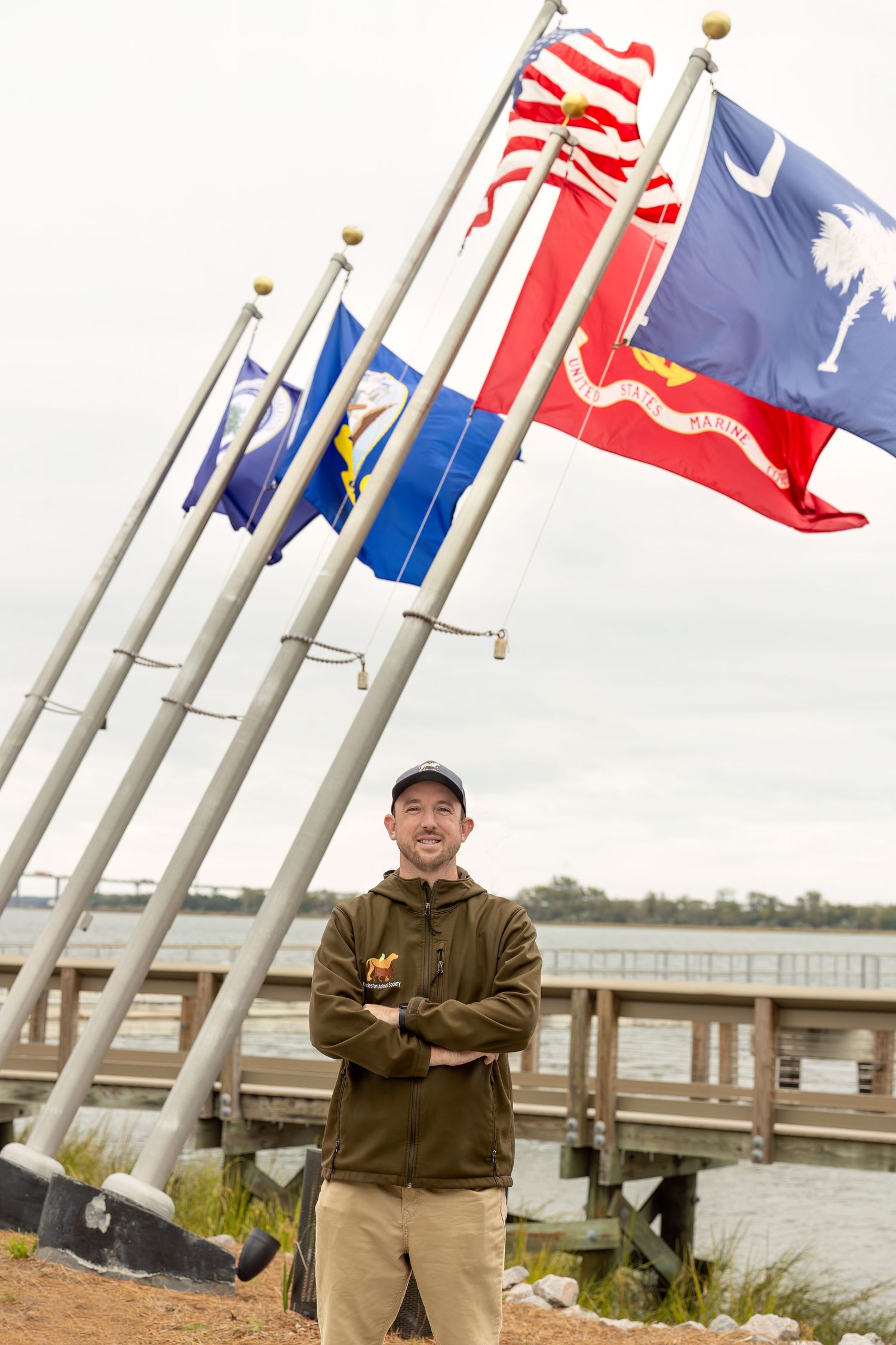 American flags wave in the wind against a cloudy sky near a waterfront with trees and a bridge visible in the distance.