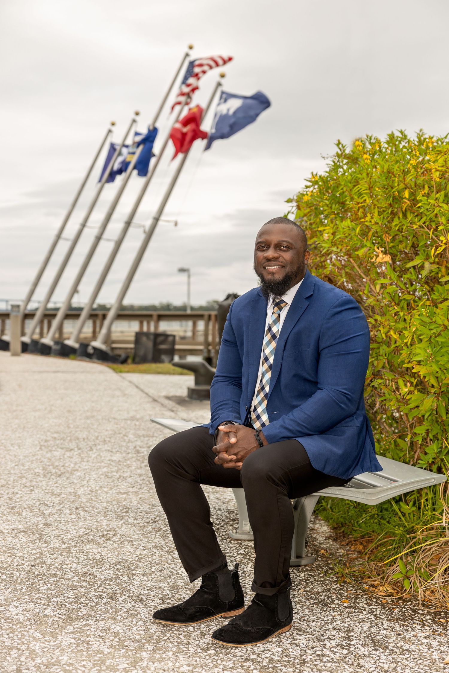 Person in blue suit sitting on bench near waterfront with flags and autumn foliage in background.