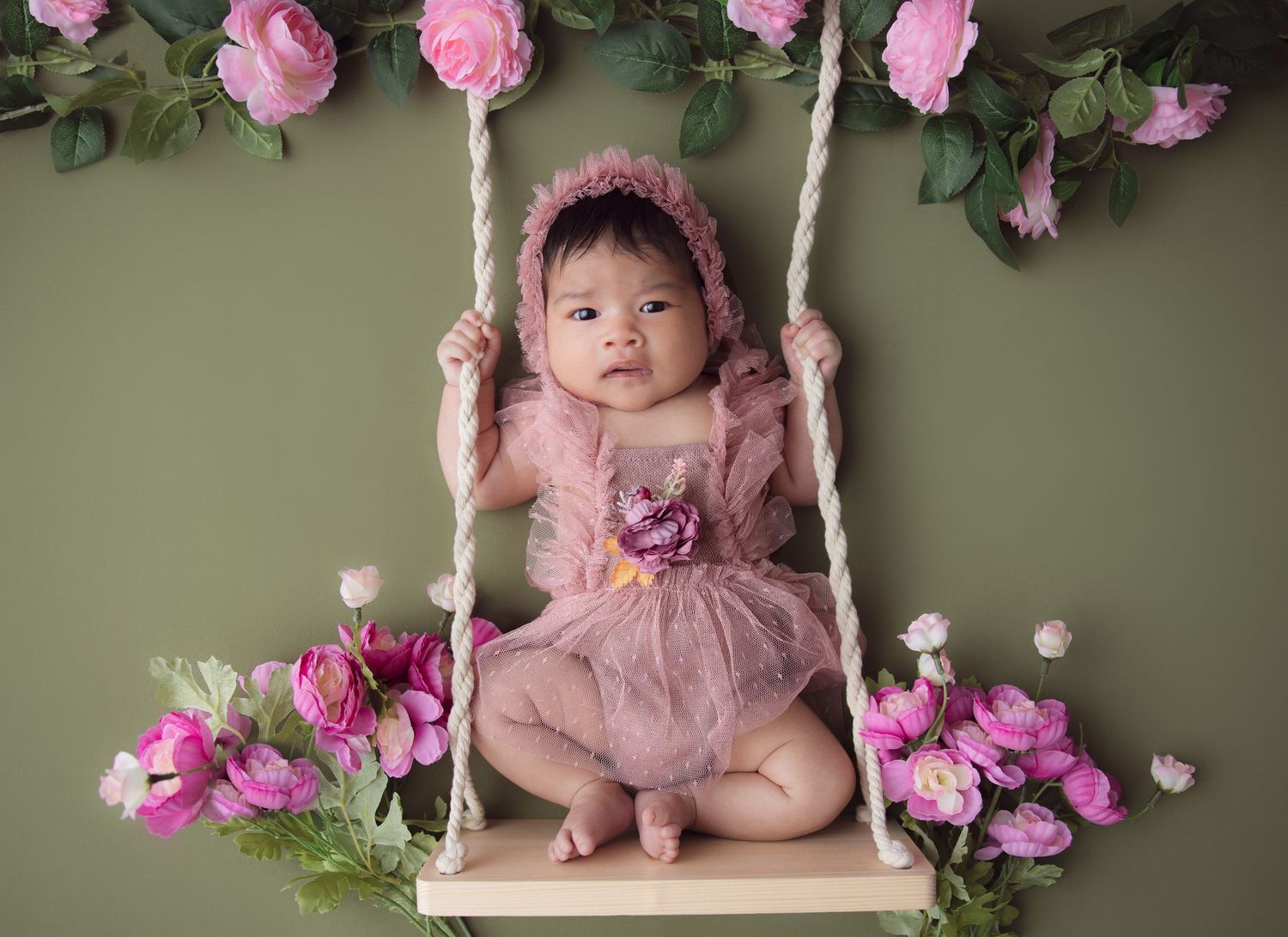 Newborn photography setup with pink roses and a wooden swing against a sage green background.