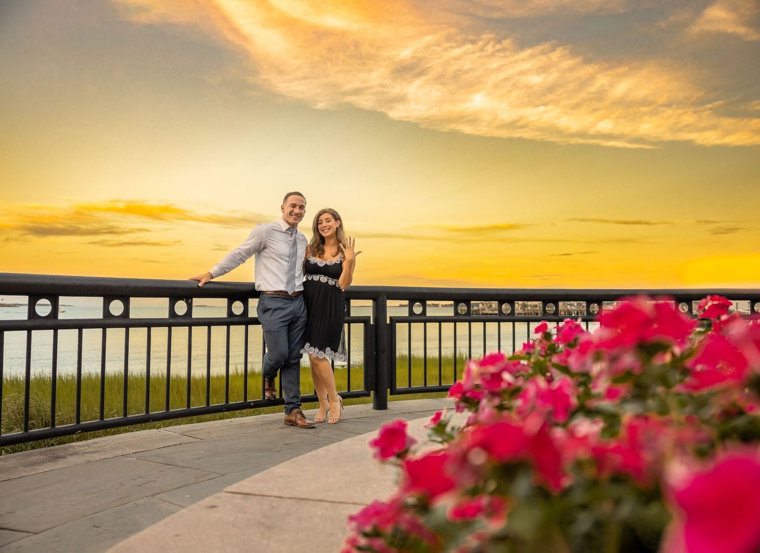 Couple shares intimate moment at sunset against railing with vibrant pink flowers and orange sky.