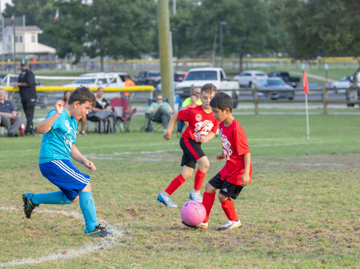 Kids playing soccer with pink ball on grass field with parked cars visible in background.