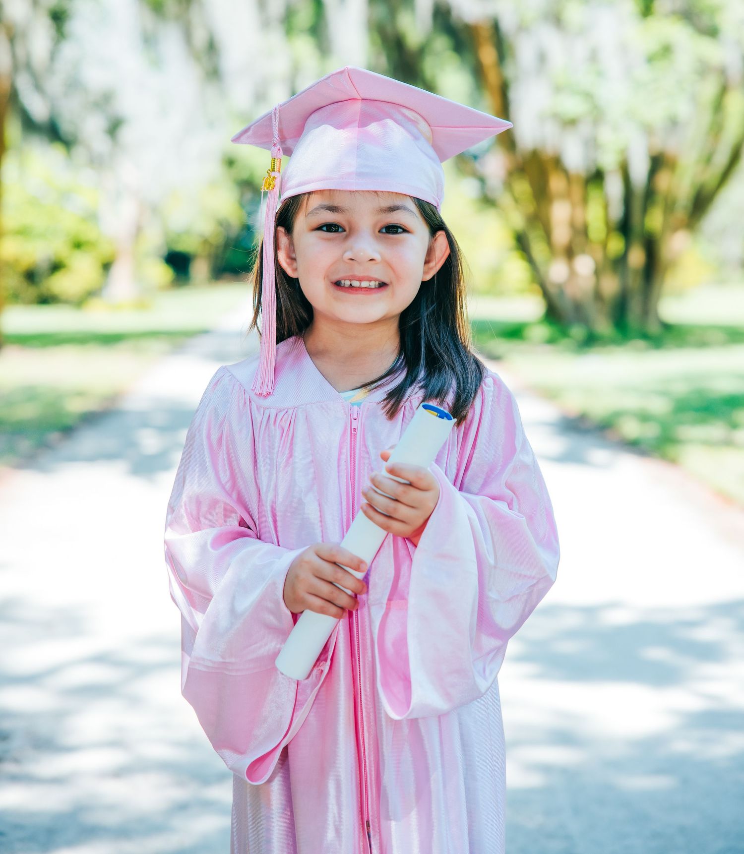 A child in pink graduation attire smiles while holding a diploma outdoors.