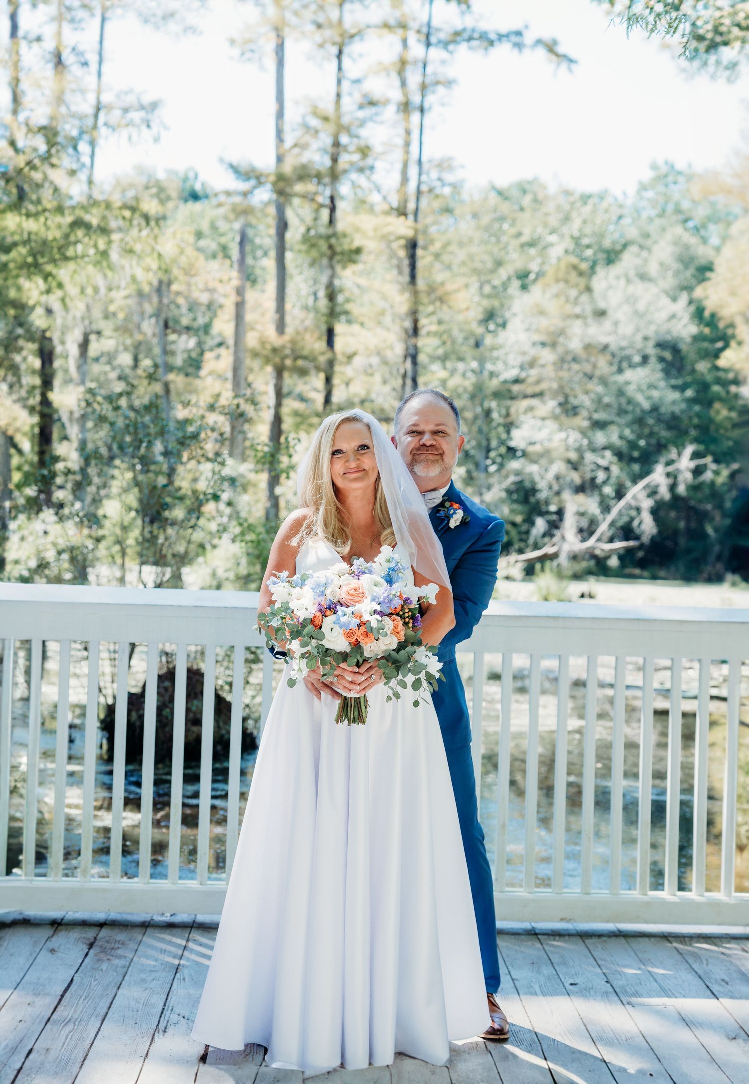 Wedding couple posing on wooden deck with white railing and pine trees in background.
