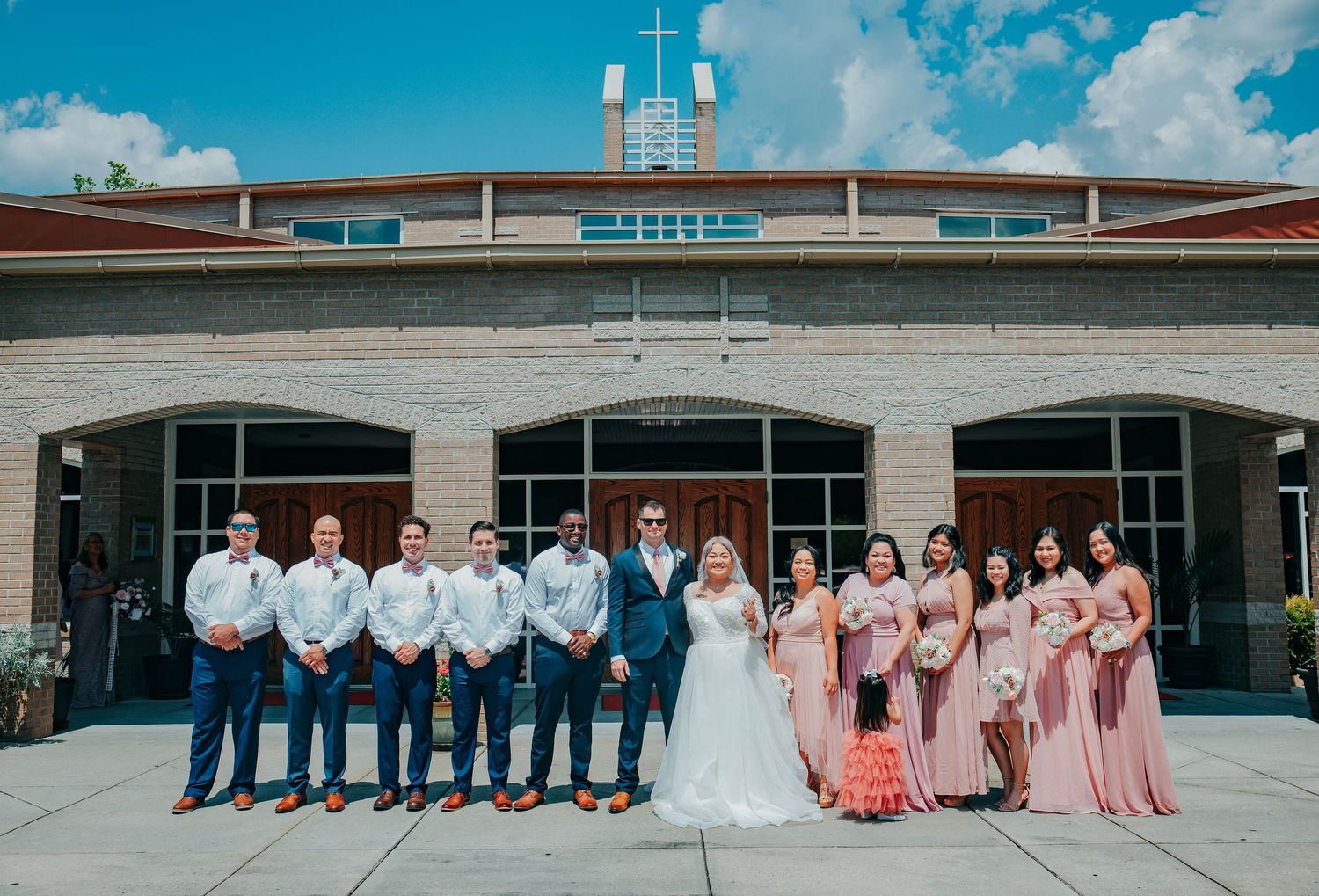 Wedding party in blue and coral attire poses in front of modern brick building under bright blue sky.