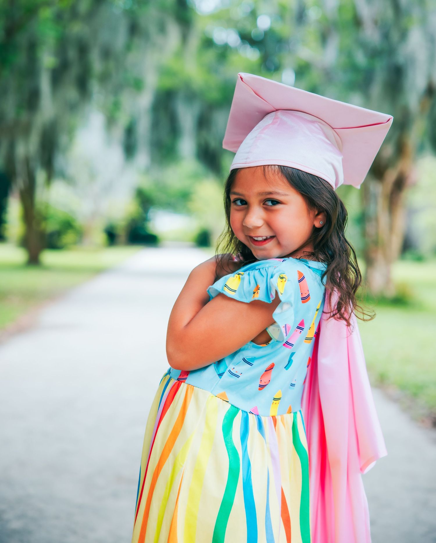 A small graduate in colorful dress twirls joyfully in a sunny outdoor setting.