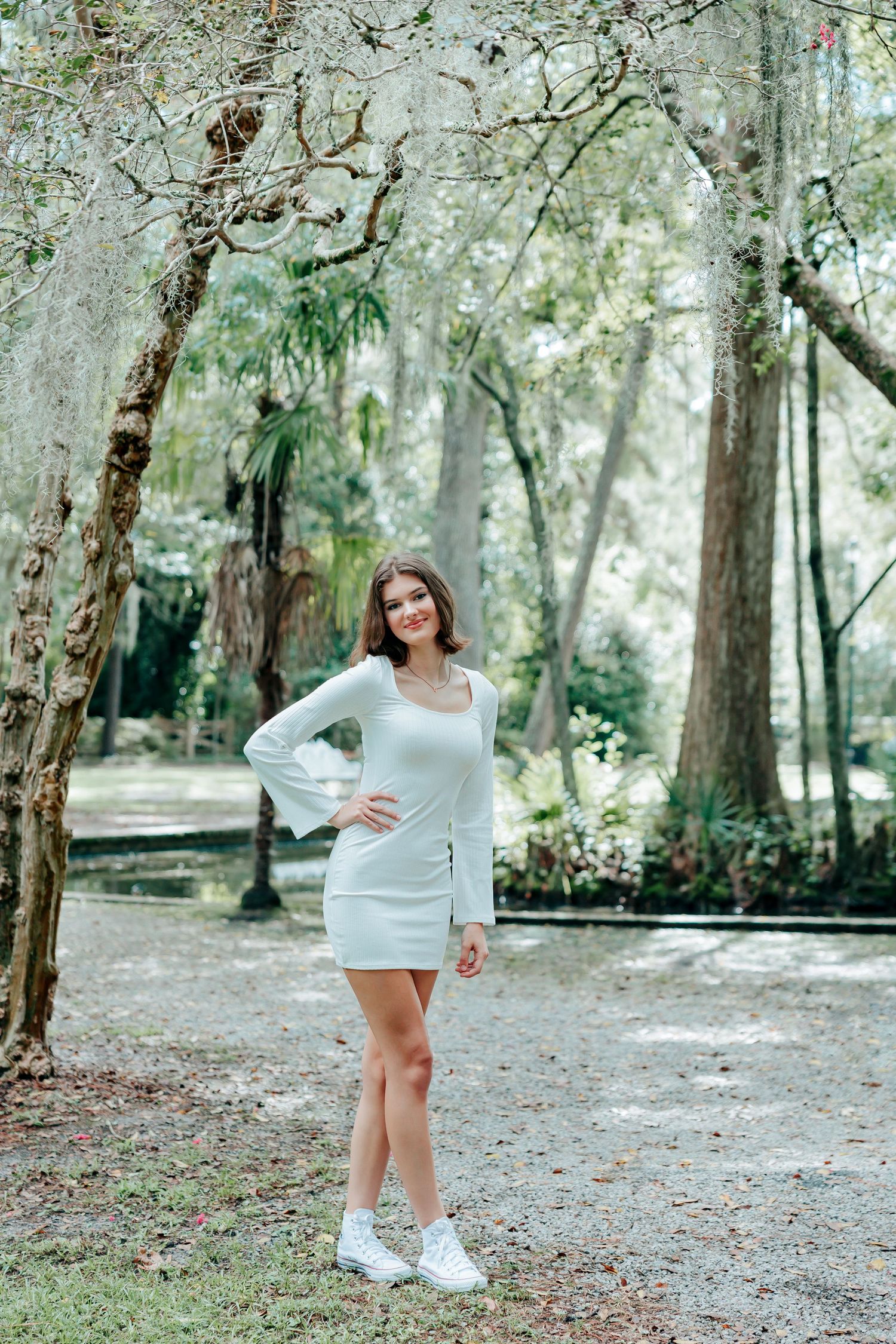 Someone wearing a white dress and sneakers poses gracefully in a wooded park setting.