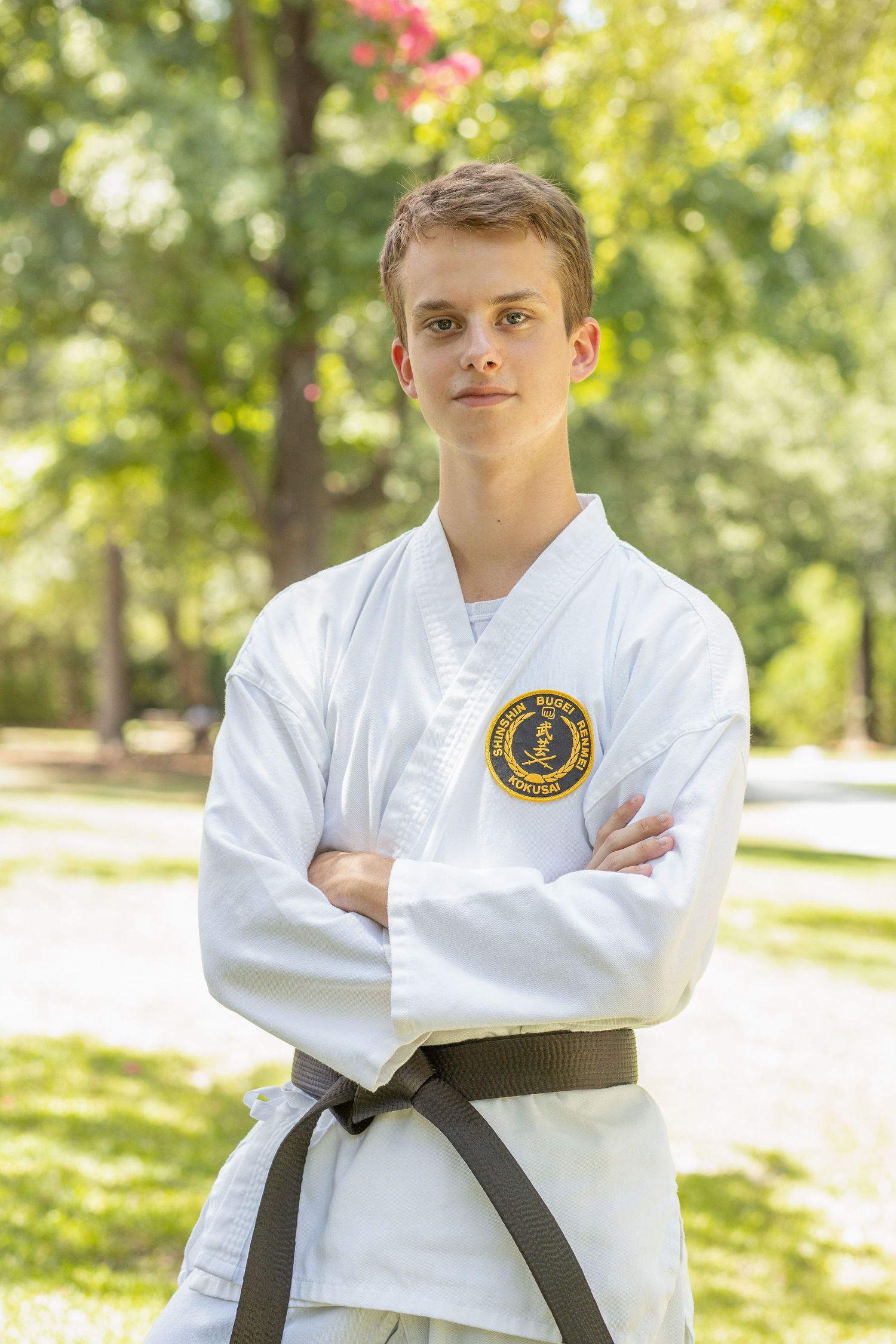 Martial arts student in white uniform with black belt poses outdoors in sunny park.