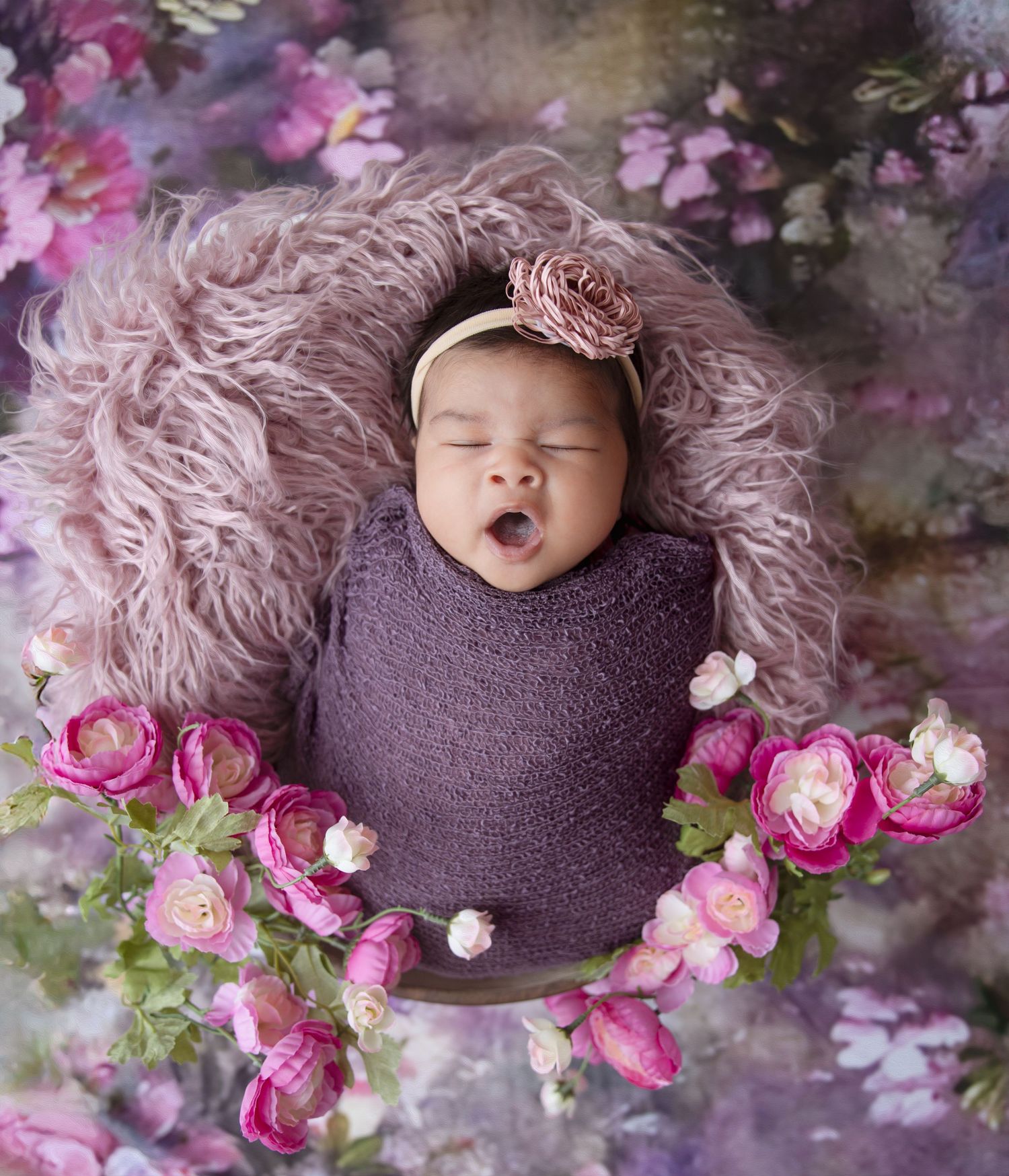 Sleeping newborn wrapped in purple fabric surrounded by pink roses in artistic arrangement.