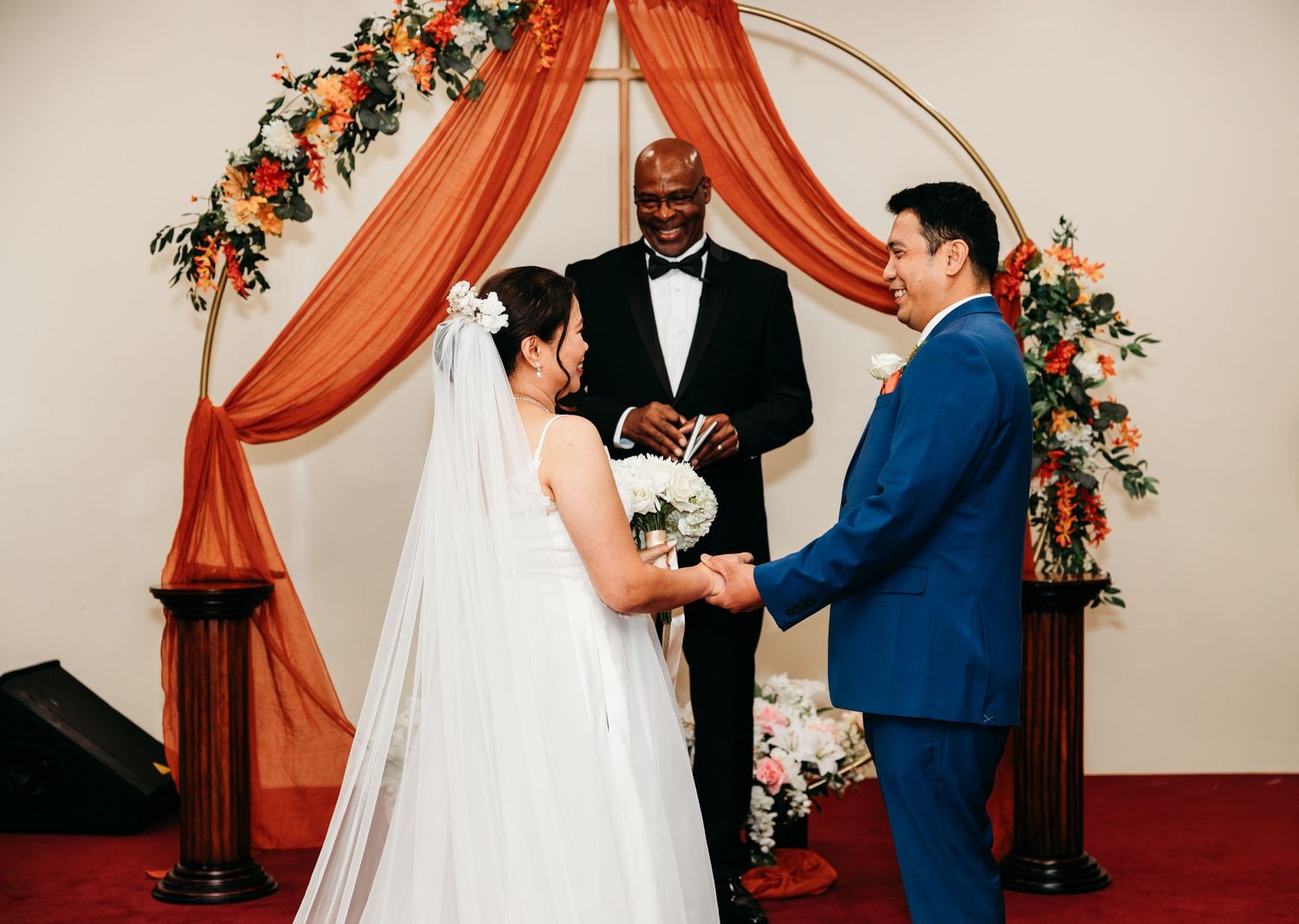 Wedding ceremony proceedings under an elegant arch with burnt orange fabric and floral details.