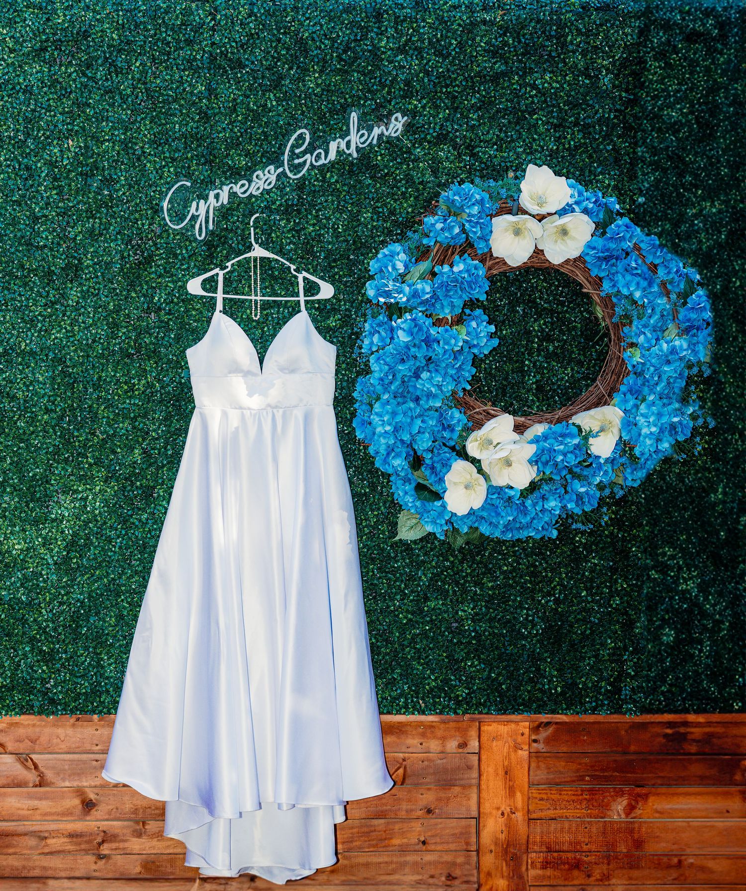 White wedding dress hanging next to a blue floral wreath against a green hedge wall backdrop.