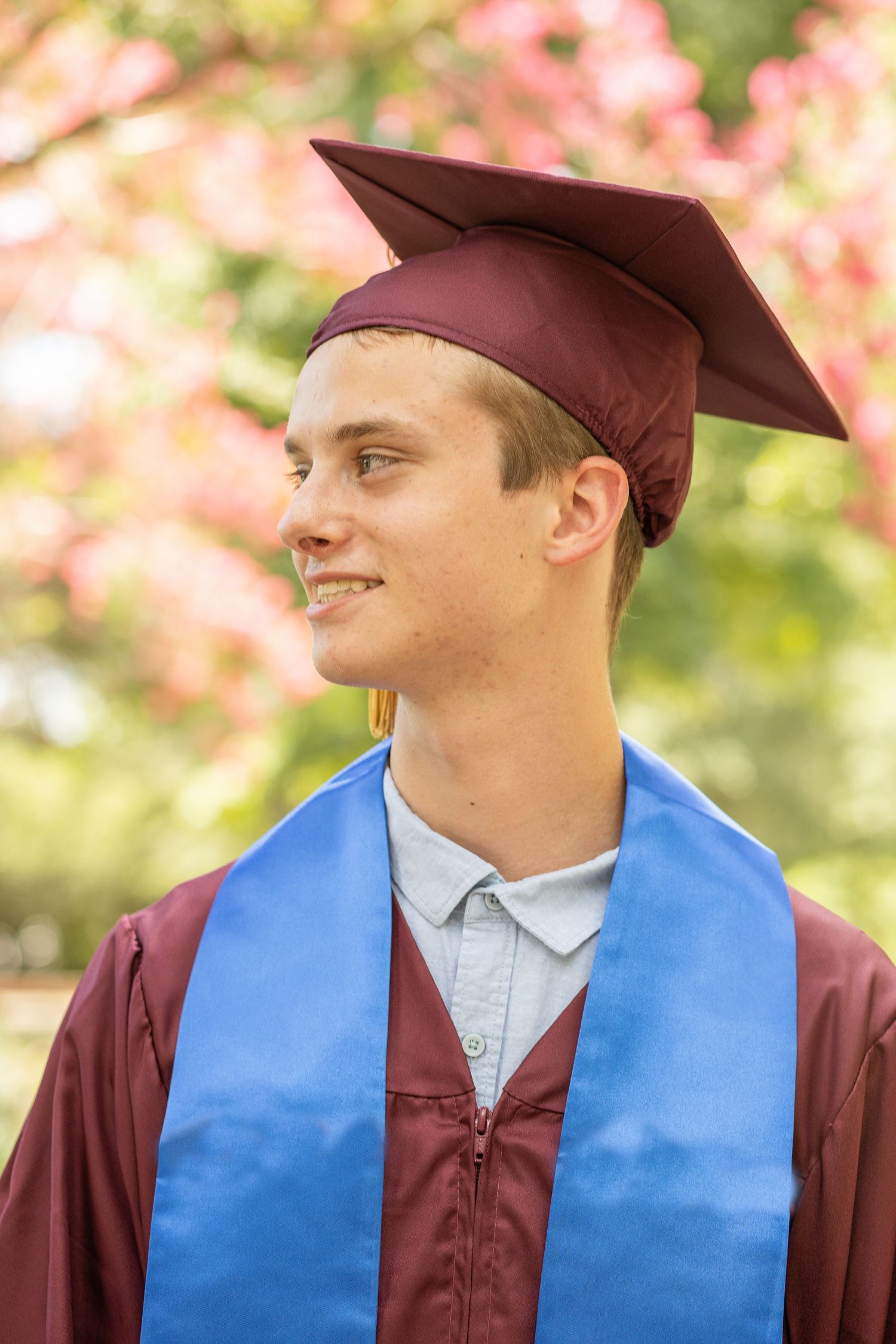 Graduate in maroon academic regalia poses against blurred pink floral background.