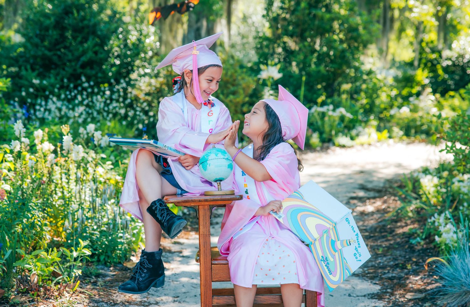A graduate sits reading on a wooden bench surrounded by garden flowers.
