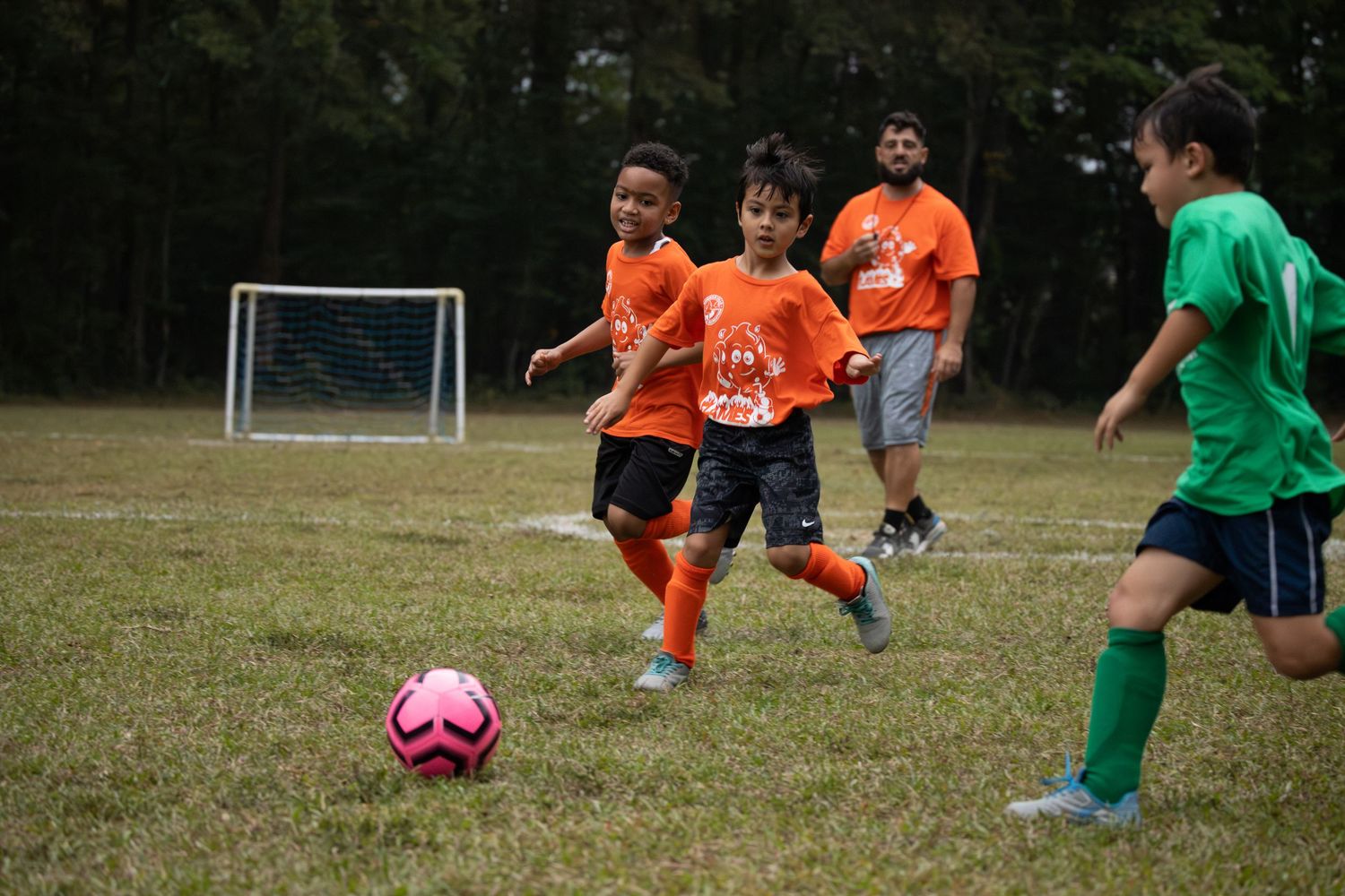 Children in orange and green uniforms playing soccer on a grass field.