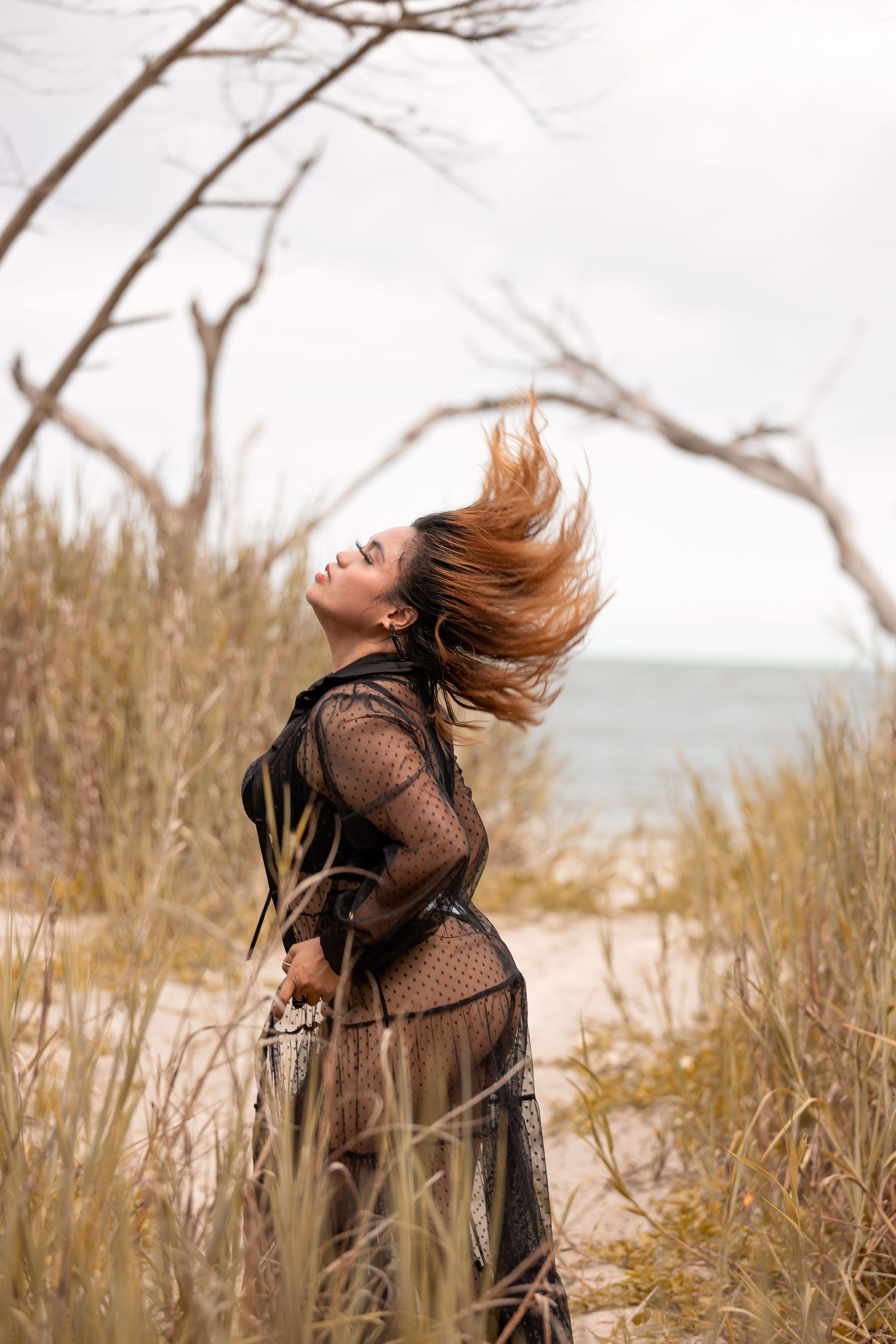 Woman with flowing hair poses dramatically in black lace dress against natural beach backdrop.