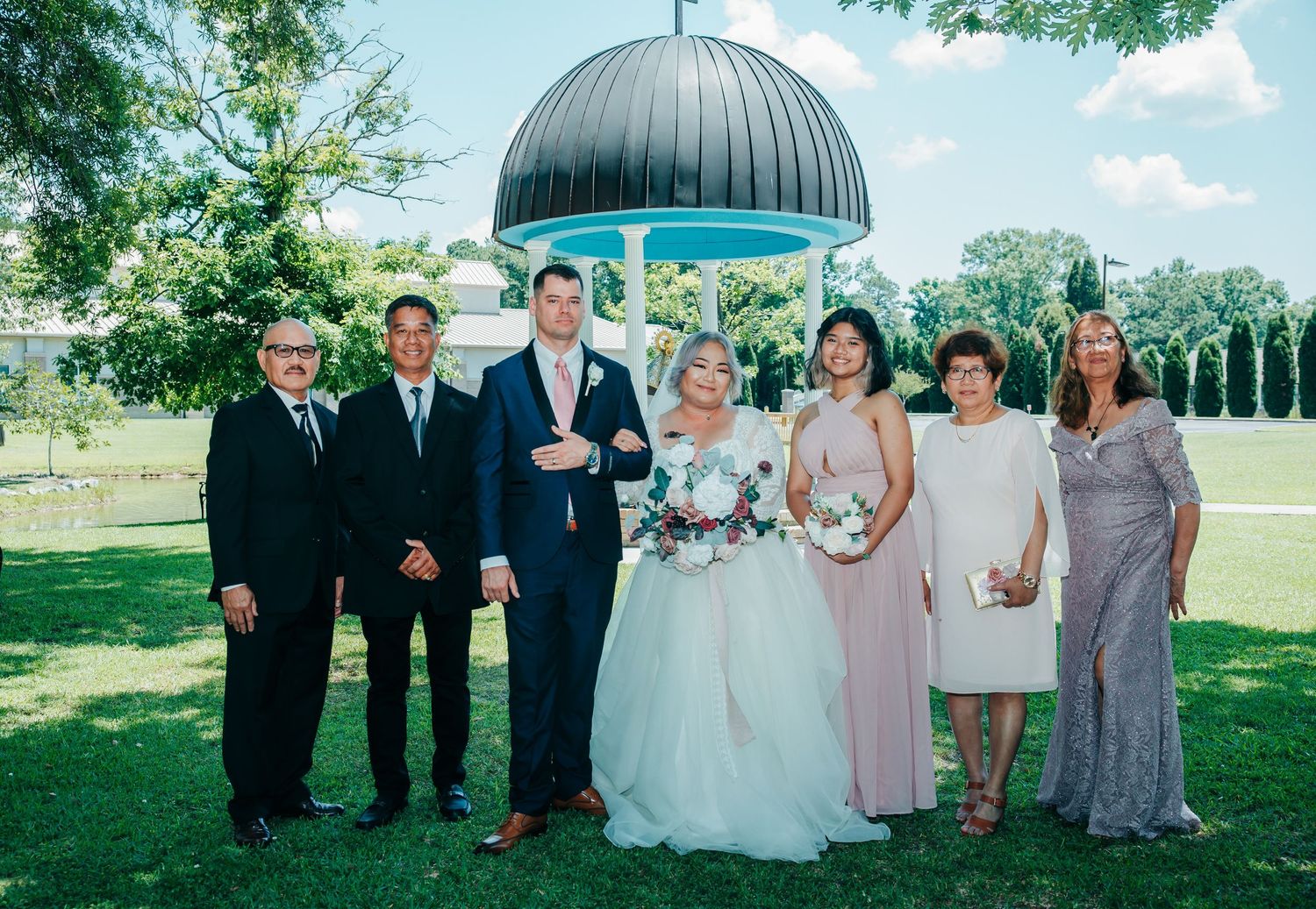 Wedding party poses outdoors on a sunny day with bridesmaids in pink dresses and groomsmen in dark suits.