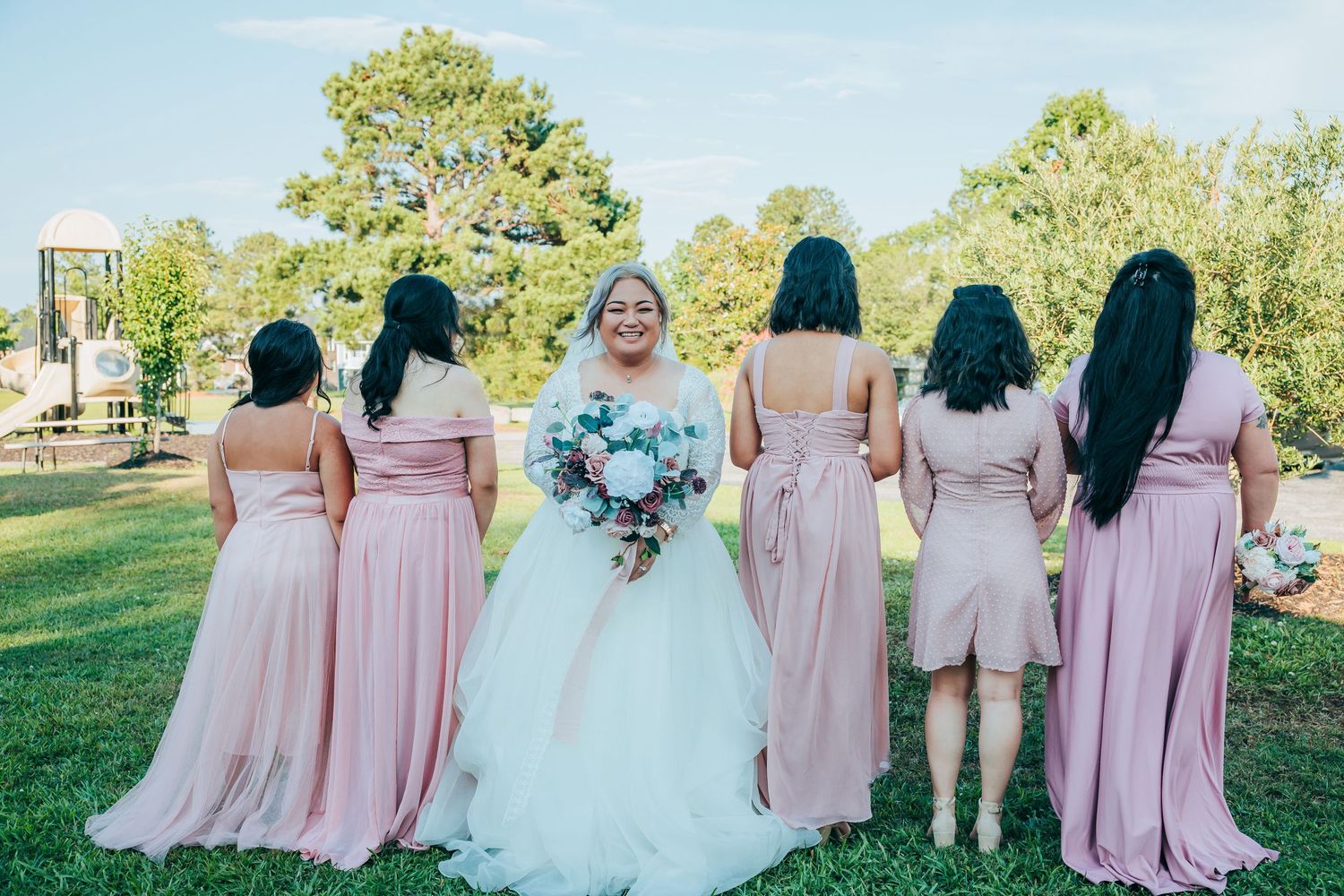 Bridesmaids in pink dresses pose with bride in white gown outdoors on sunny day.