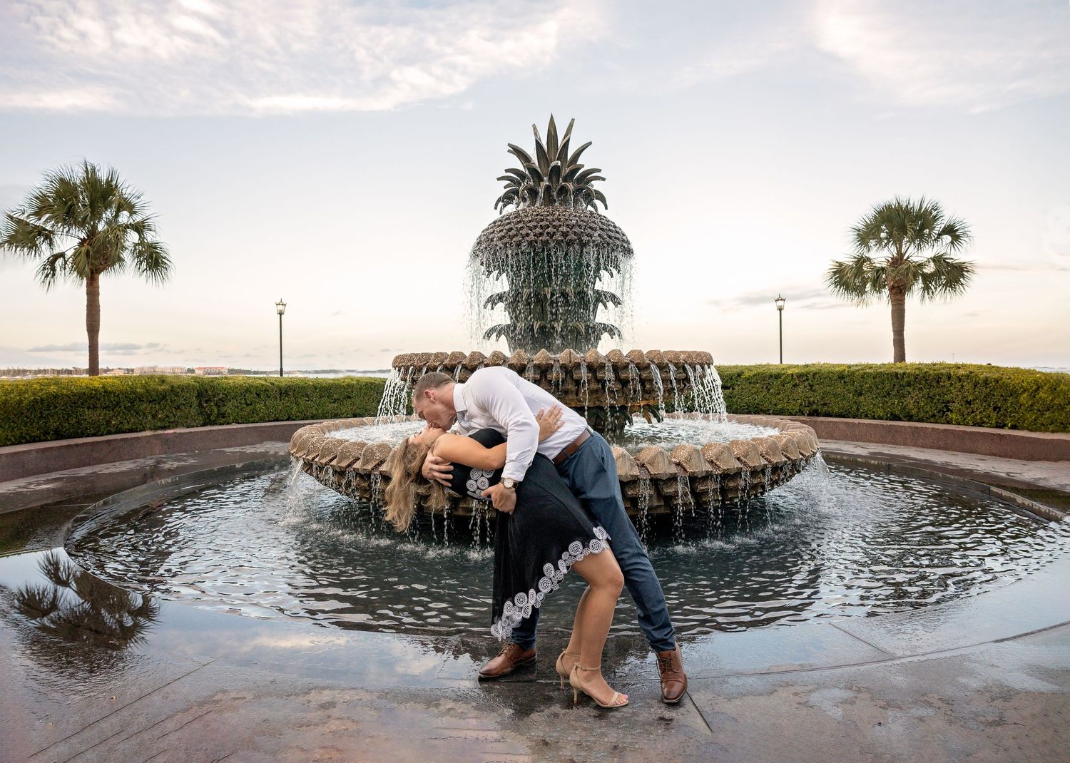 Couple poses playfully by ornate pineapple fountain during sunset photoshoot.